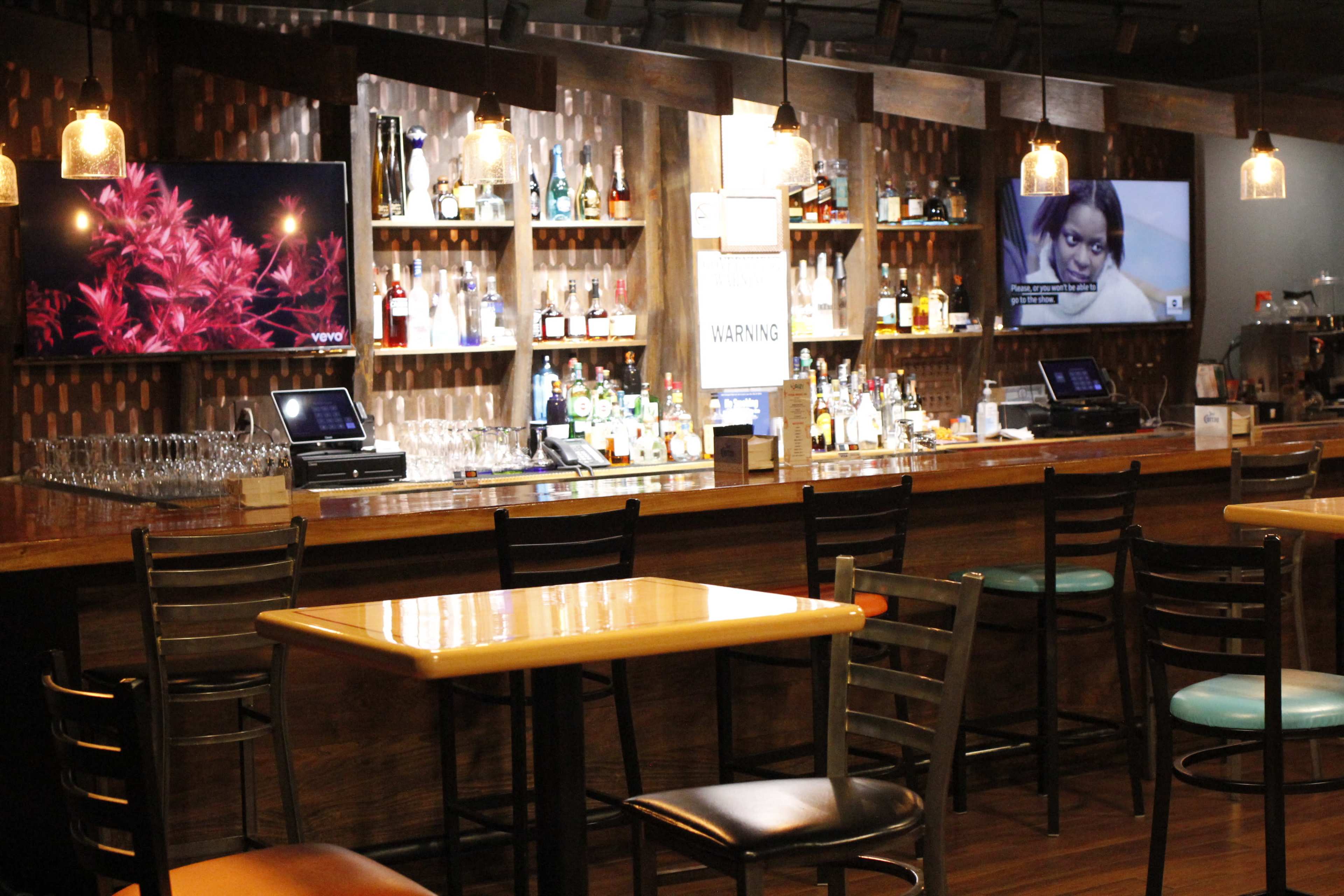 A bar with a wooden counter, multiple shelves of liquor bottles, and two televisions mounted on the walls.