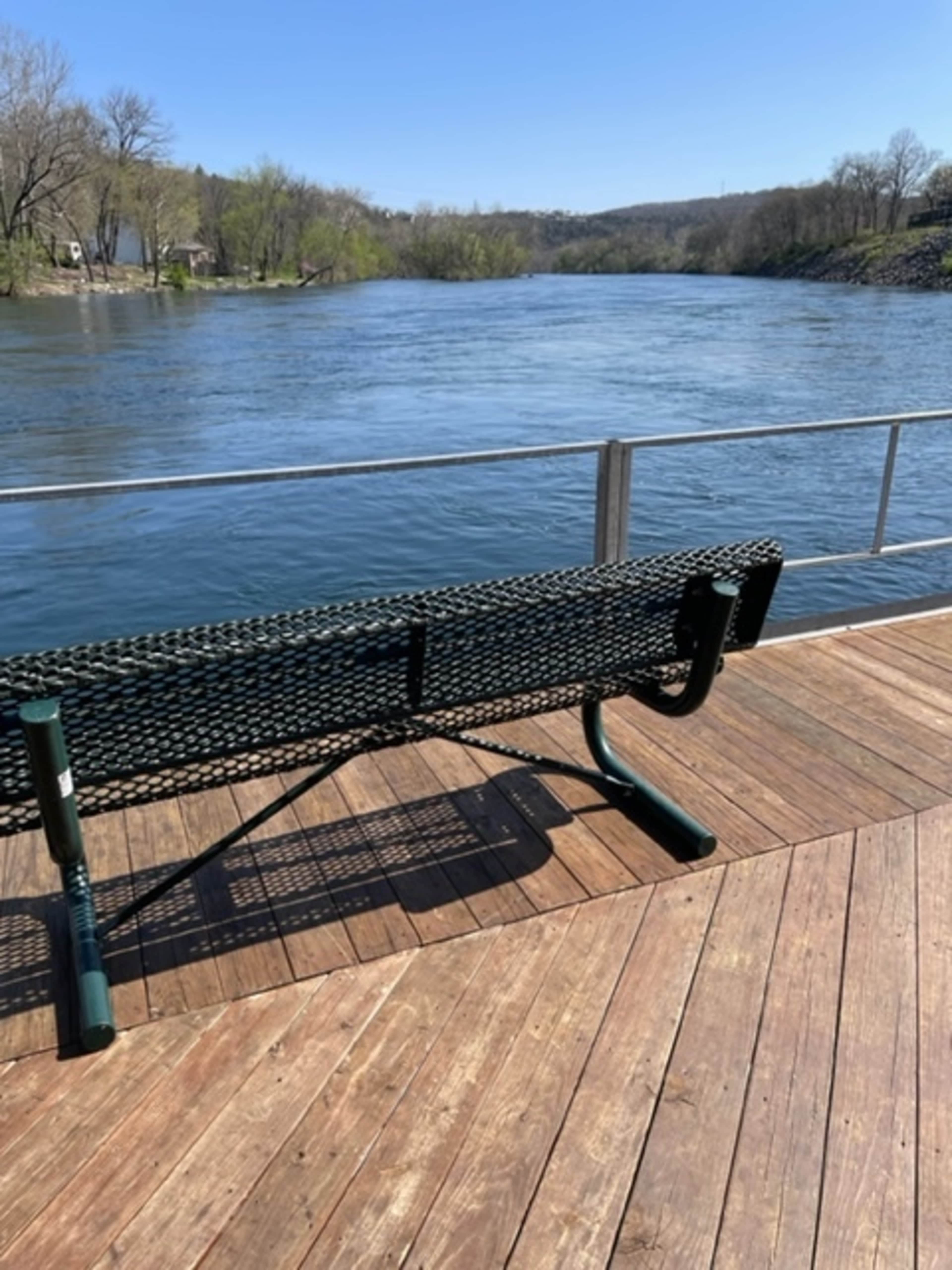 A green bench is positioned on a wooden platform overlooking a calm river under a clear sky.