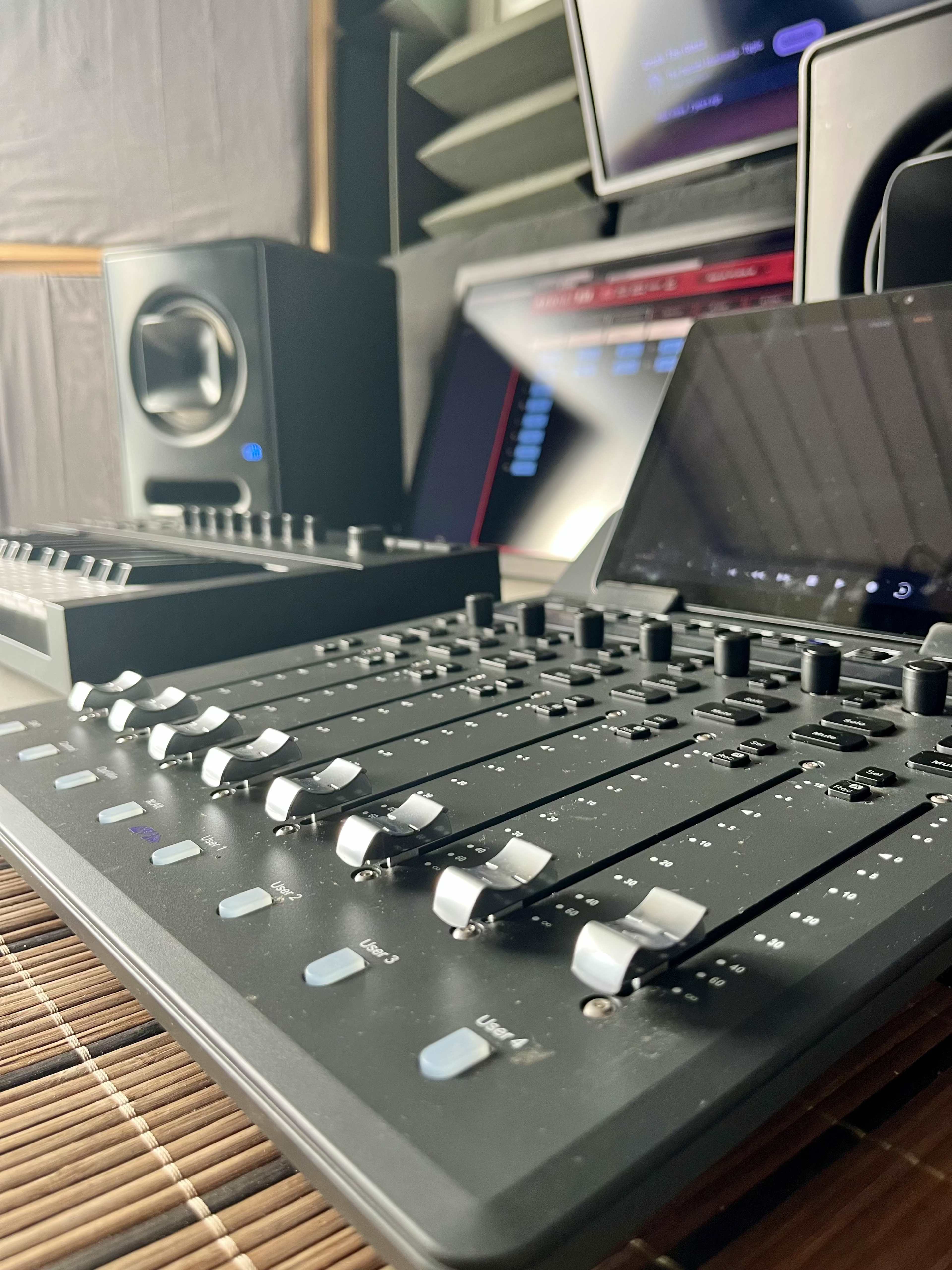 The image shows a close-up of a digital audio mixing console with faders and controls, set in a music production studio alongside speakers and computer monitors.