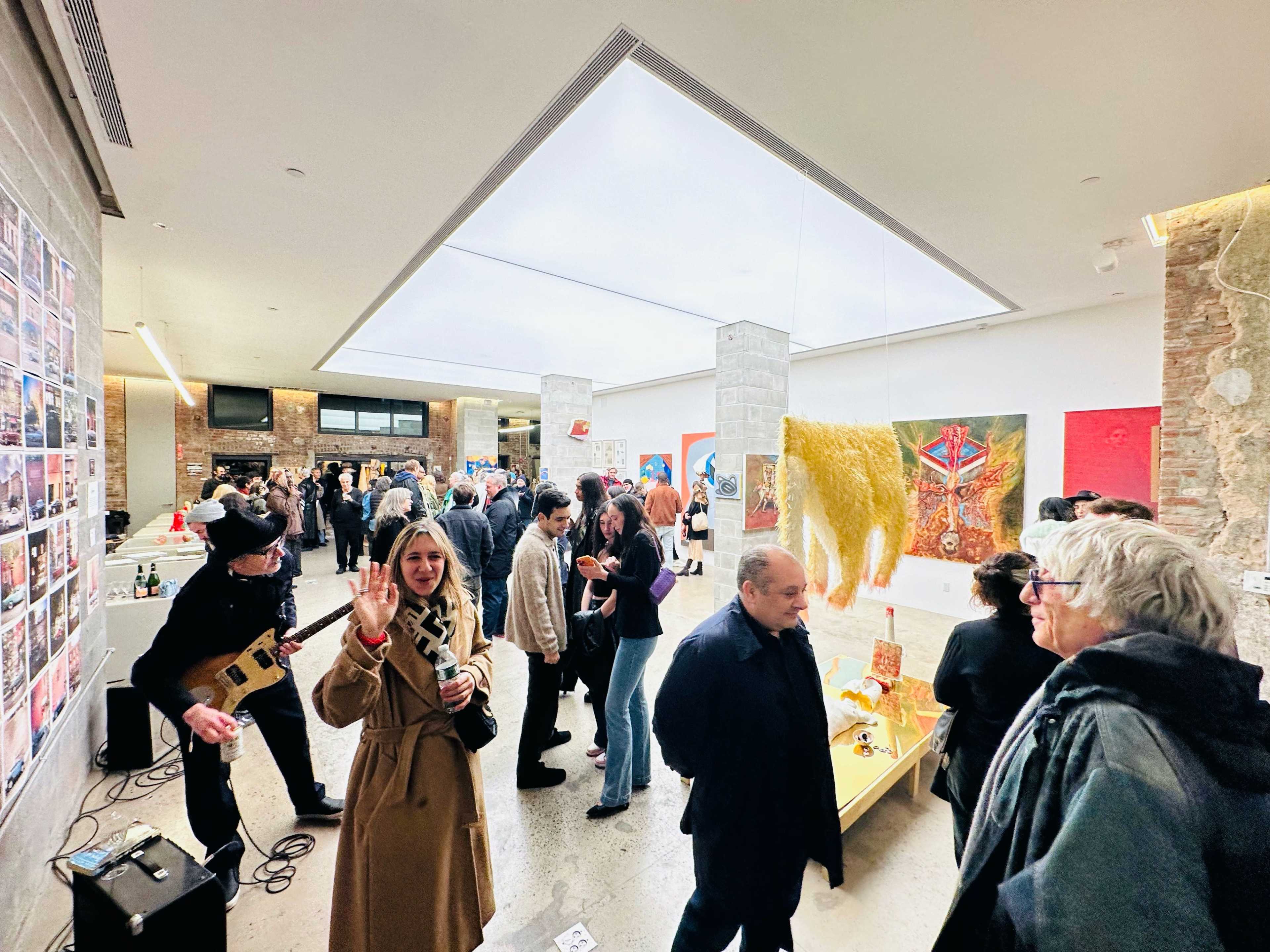 A crowd of people mingles at an art gallery opening, surrounded by colorful artwork displayed on the walls and tables.
