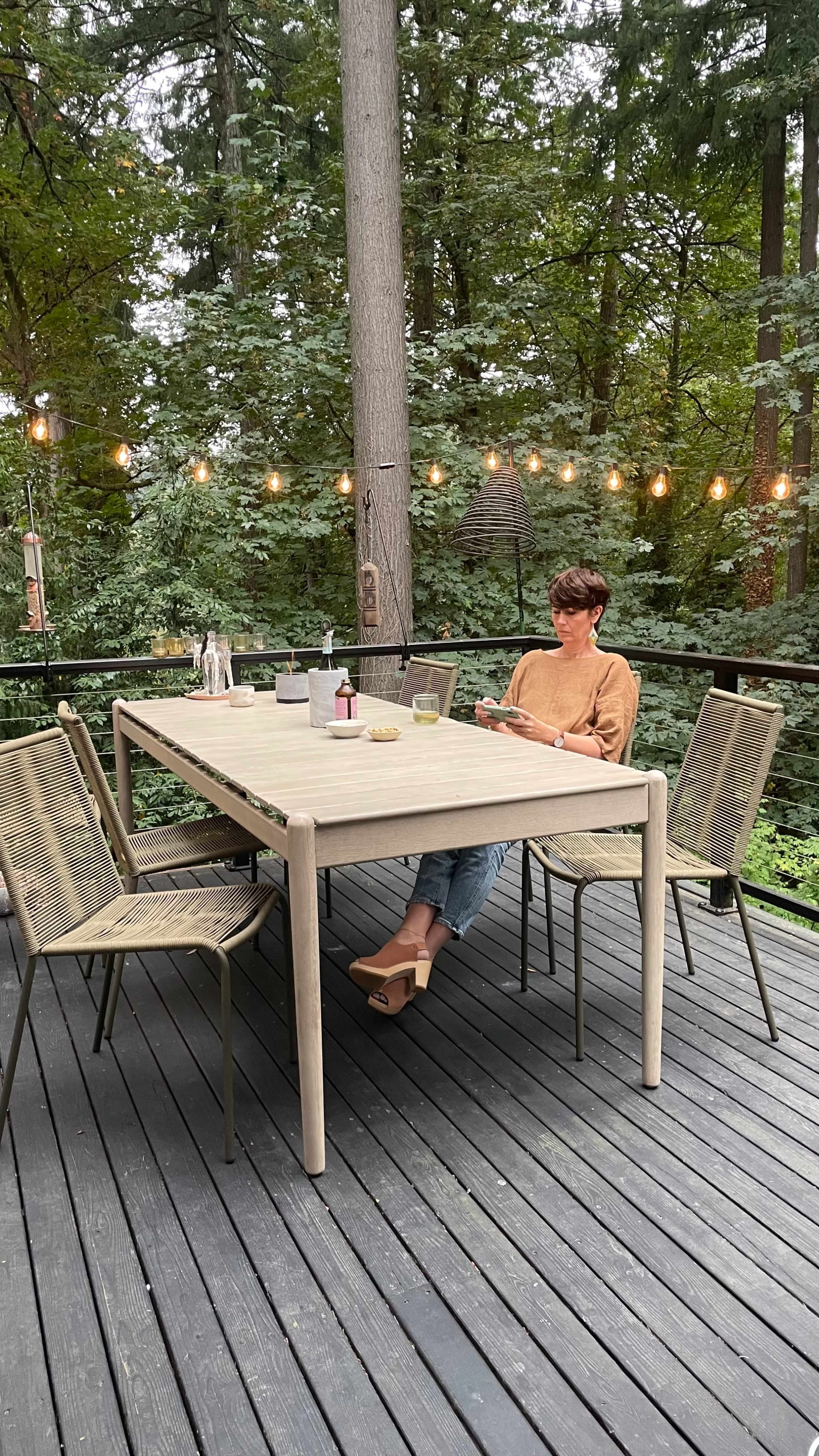 A woman sits at a wooden table on a deck surrounded by trees, with string lights hanging above and beverages and cookies on the table.