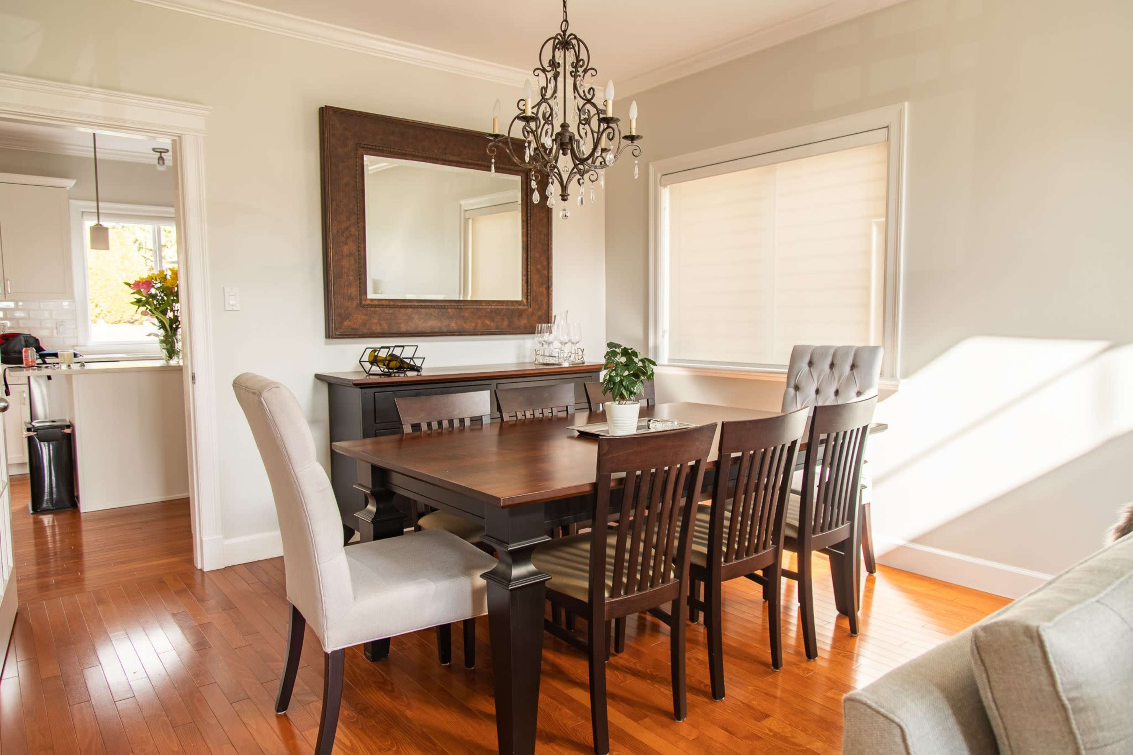 A dining room features a large wooden table surrounded by chairs, with a chandelier hanging above and a mirror on the wall.