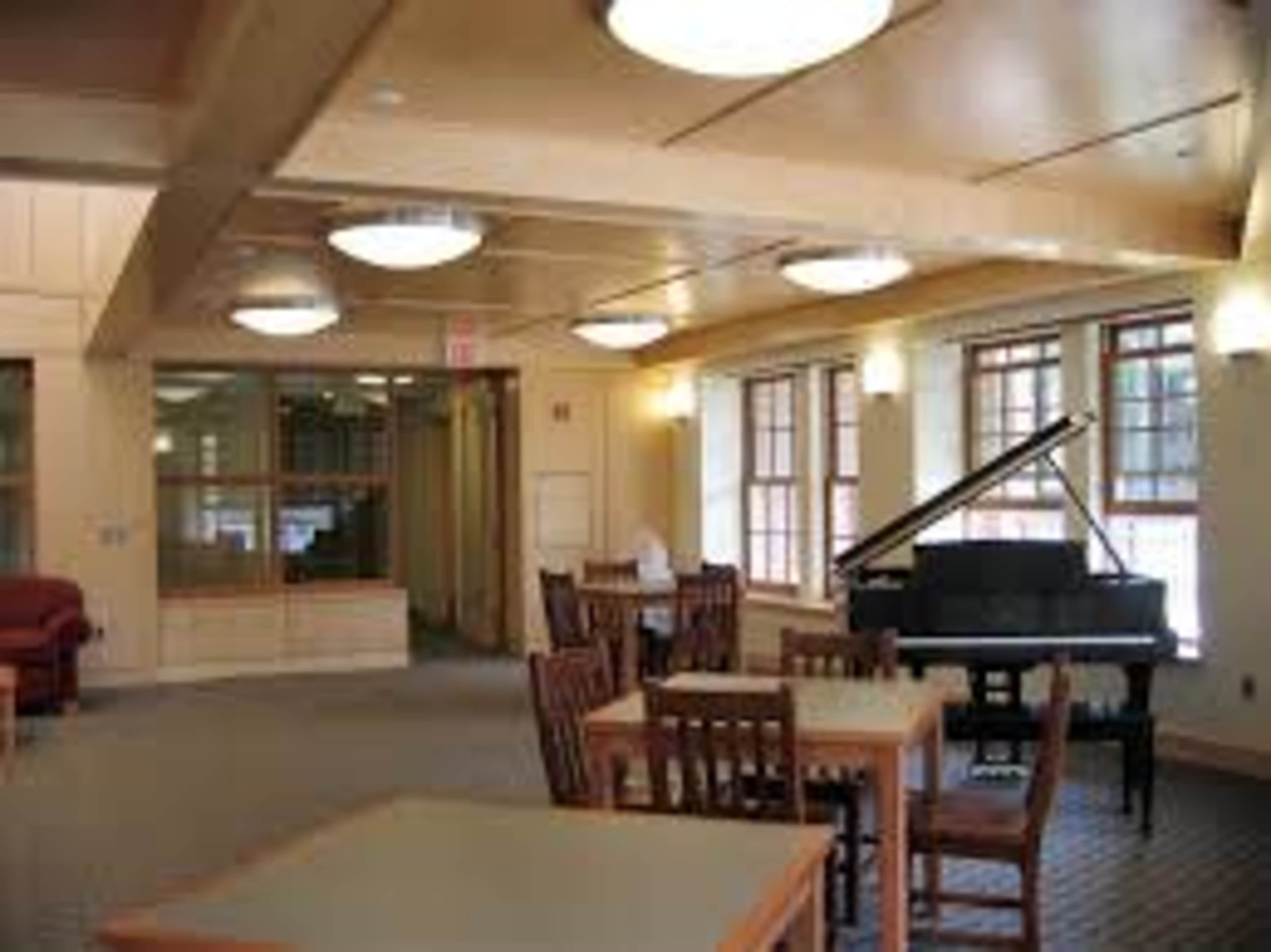 The image shows a spacious room featuring a grand piano, tables, and wooden chairs, with large windows allowing natural light to enter.