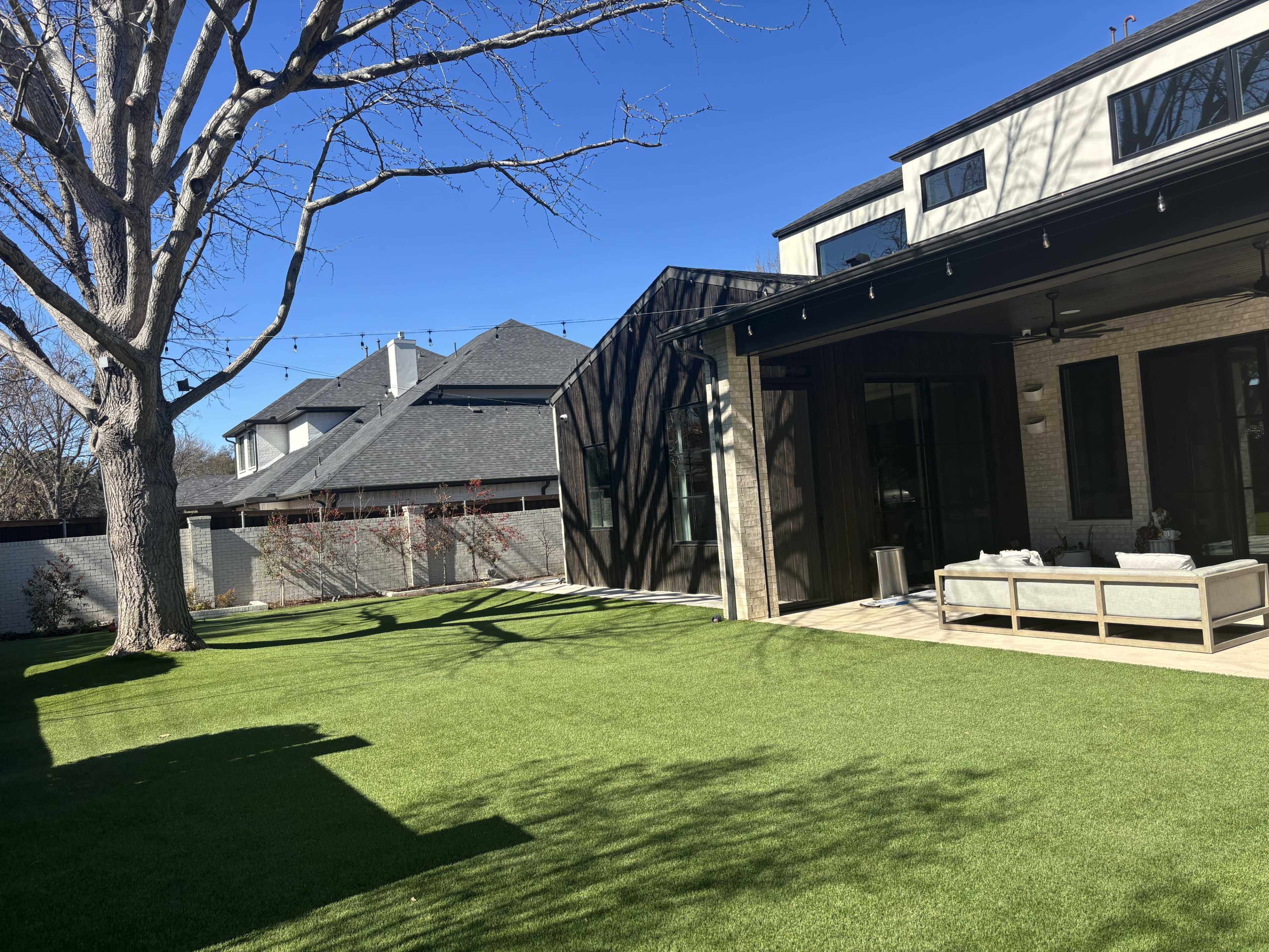 The image shows a well-maintained lawn with a modern home featuring a glass wall, and a tree in the foreground under a clear blue sky.