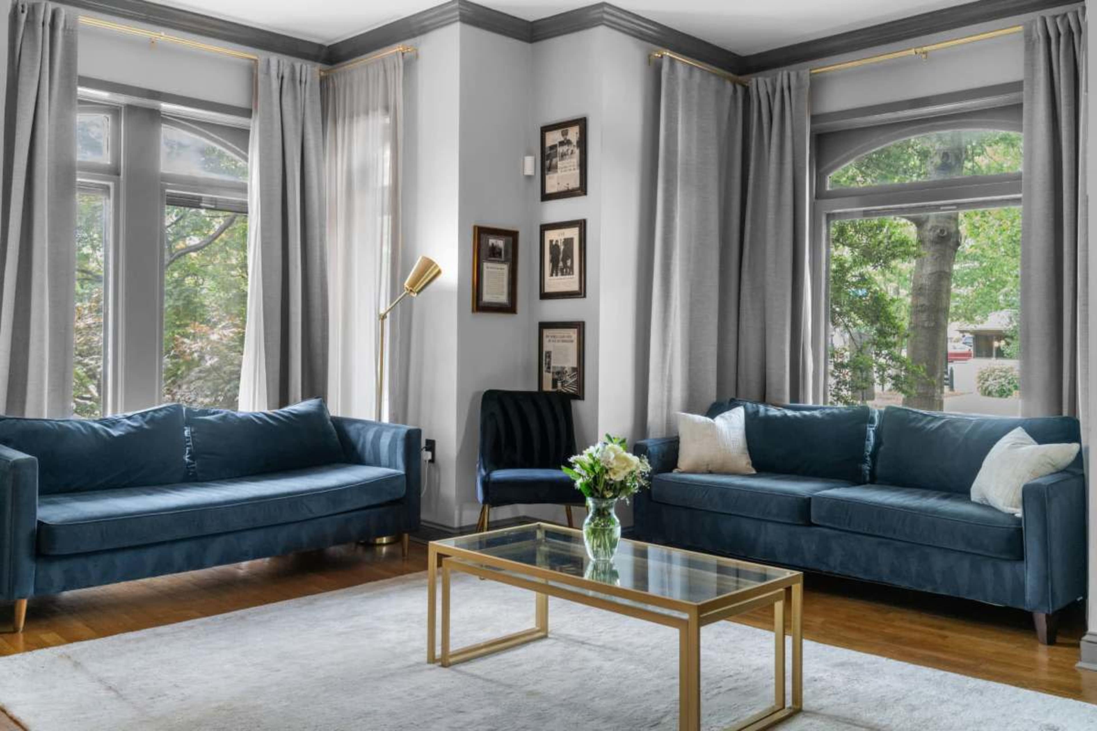 The image features a living room with two blue sofas and a glass coffee table, surrounded by light curtains and framed artwork on the walls.
