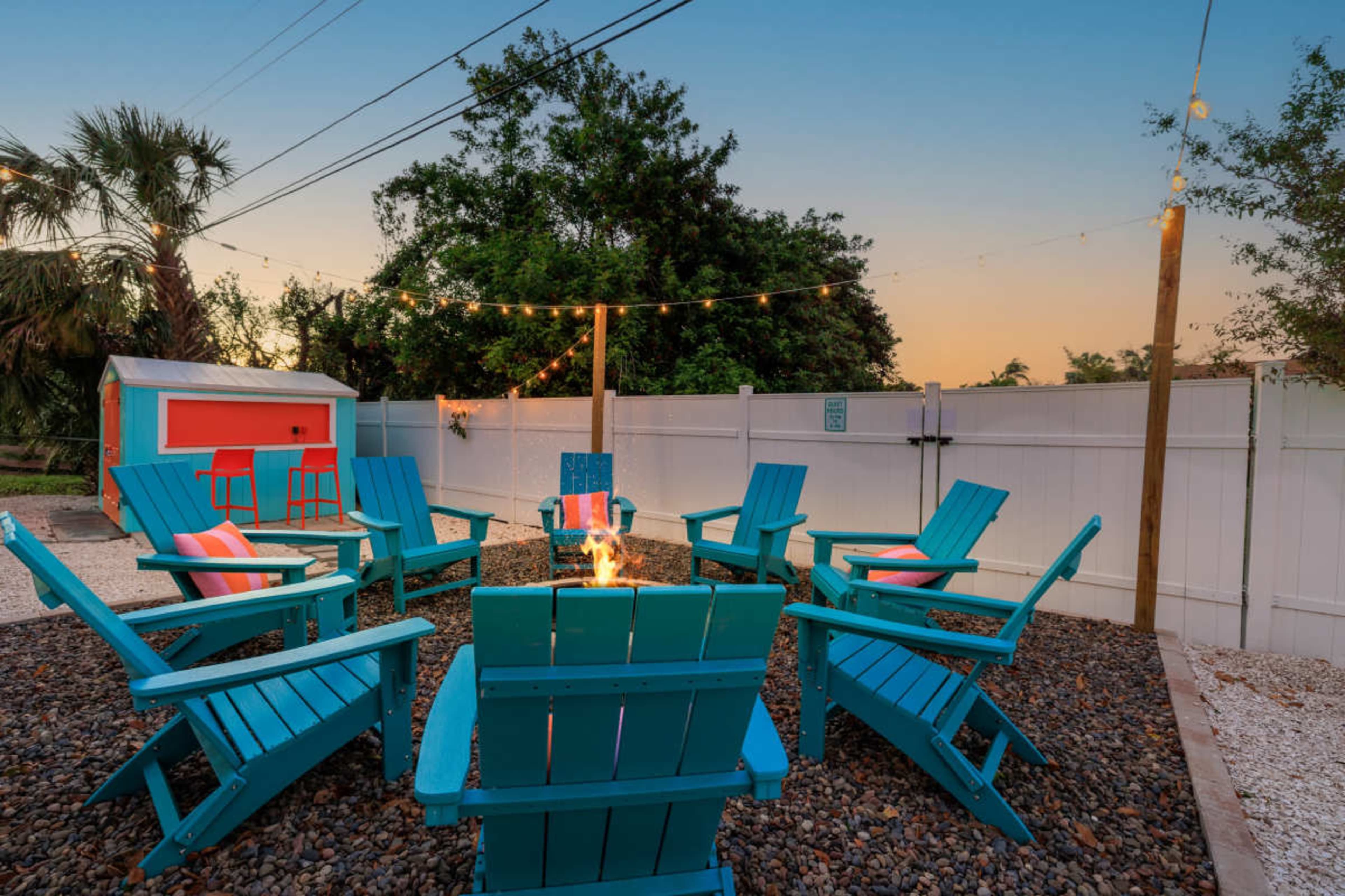 A circular arrangement of bright blue Adirondack chairs surrounds a fire pit in a backyard, with string lights overhead and trees in the background.
