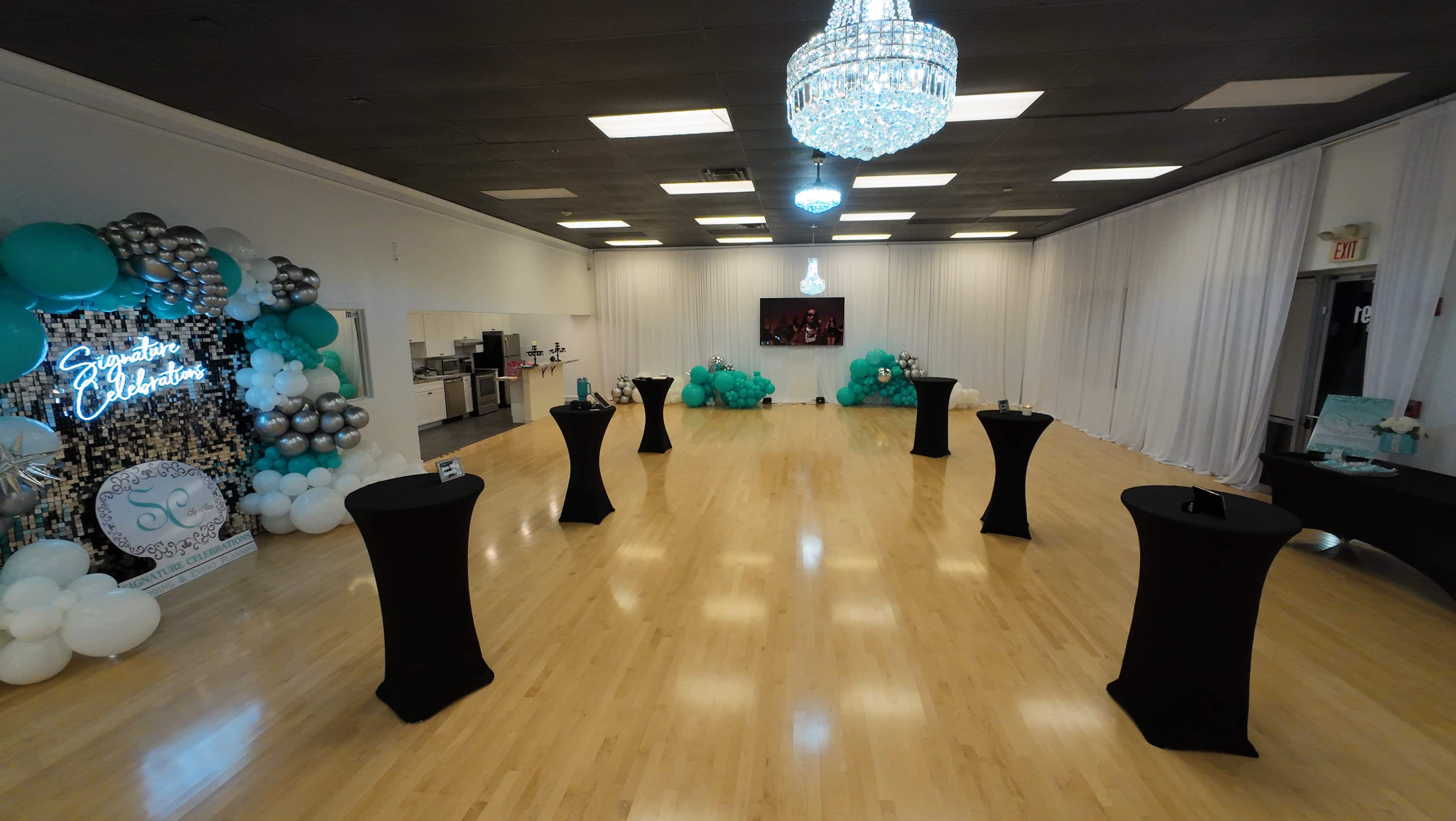 An event space featuring several round tables with black covers, turquoise decorations, and a large chandelier overhead.
