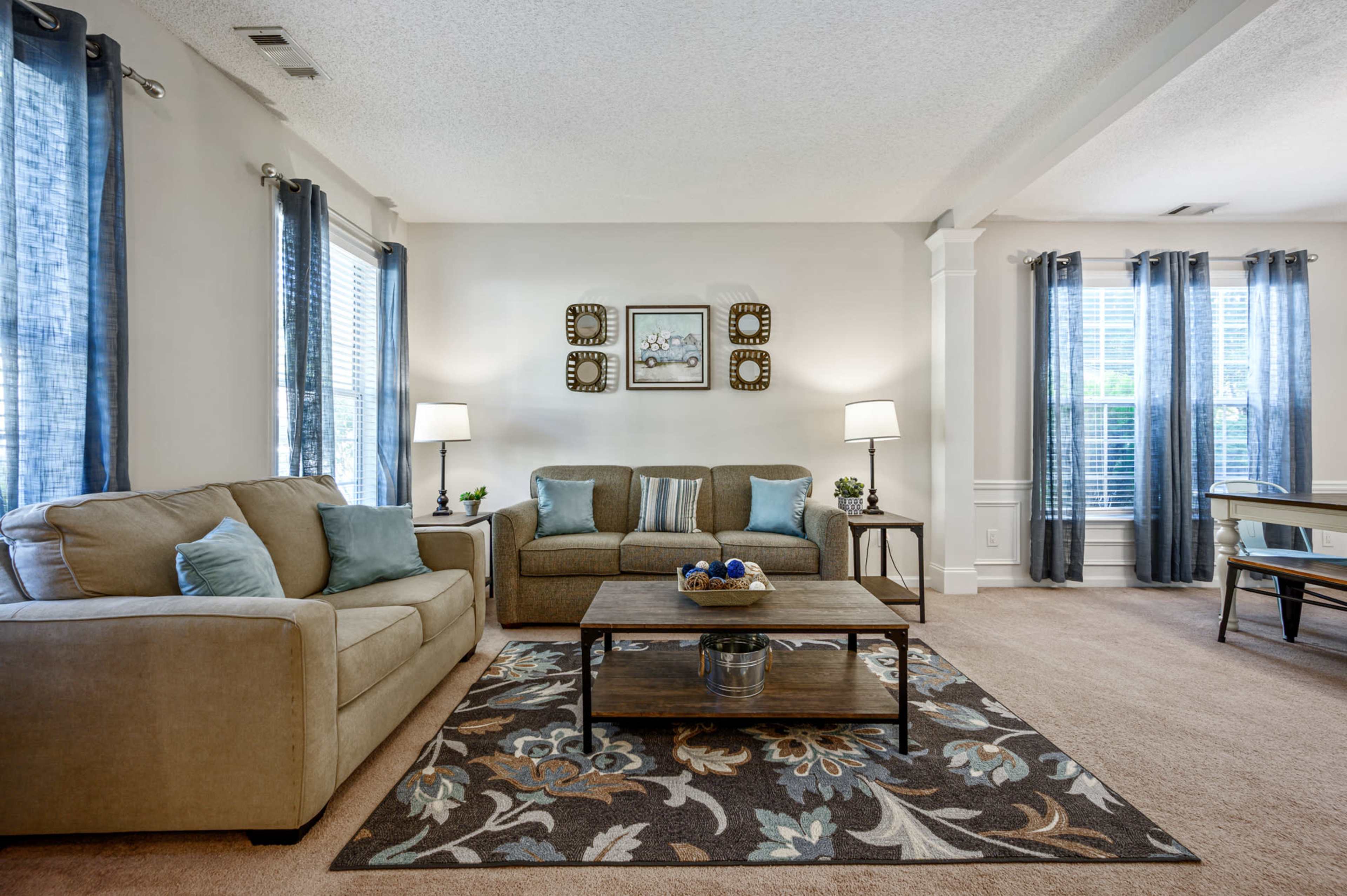 A cozy living room features a beige couch and loveseat, a wooden coffee table, and large windows with blue curtains.