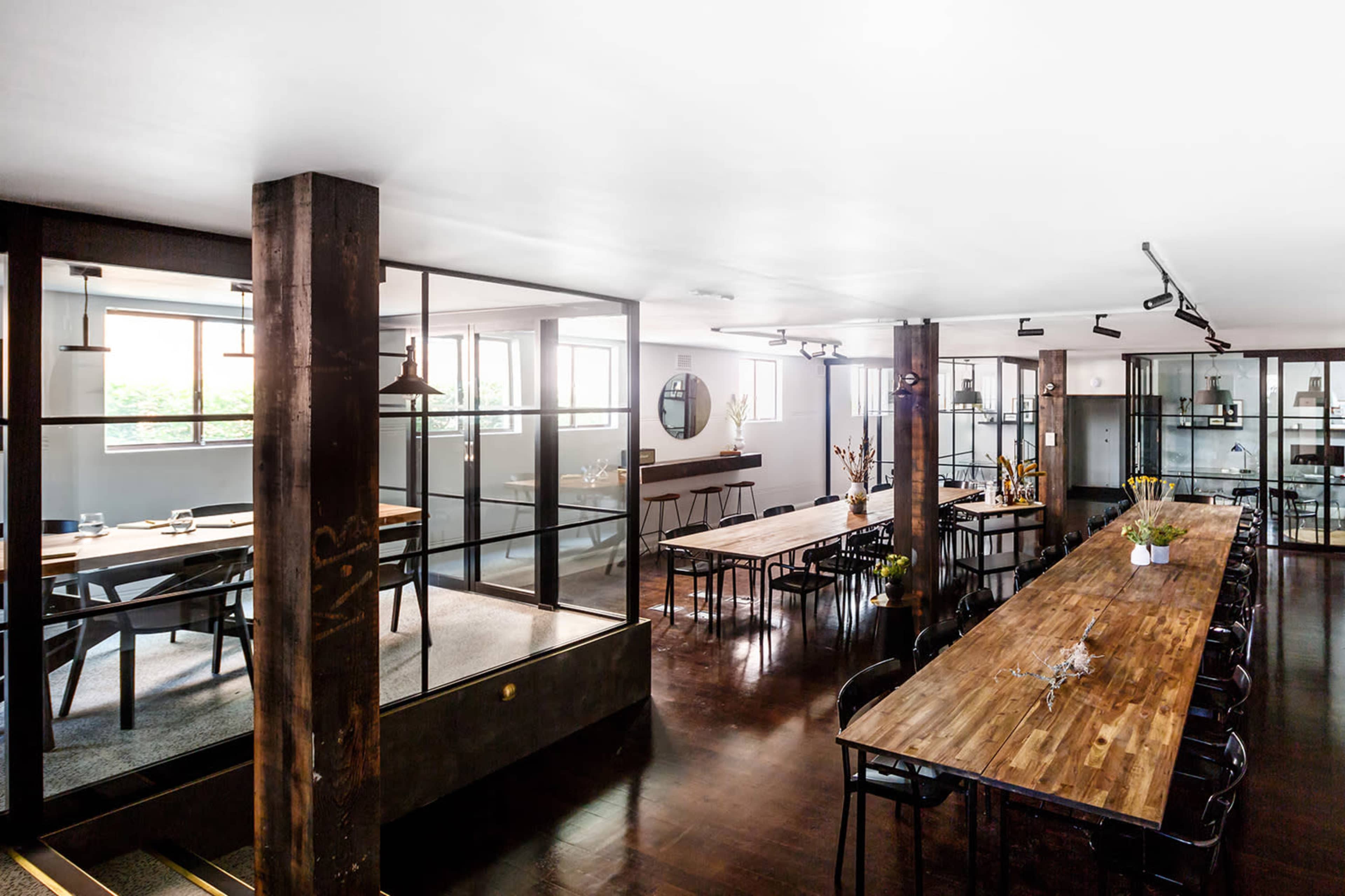 The image shows a modern office space featuring wooden tables, glass partitions, and a variety of seating arrangements.