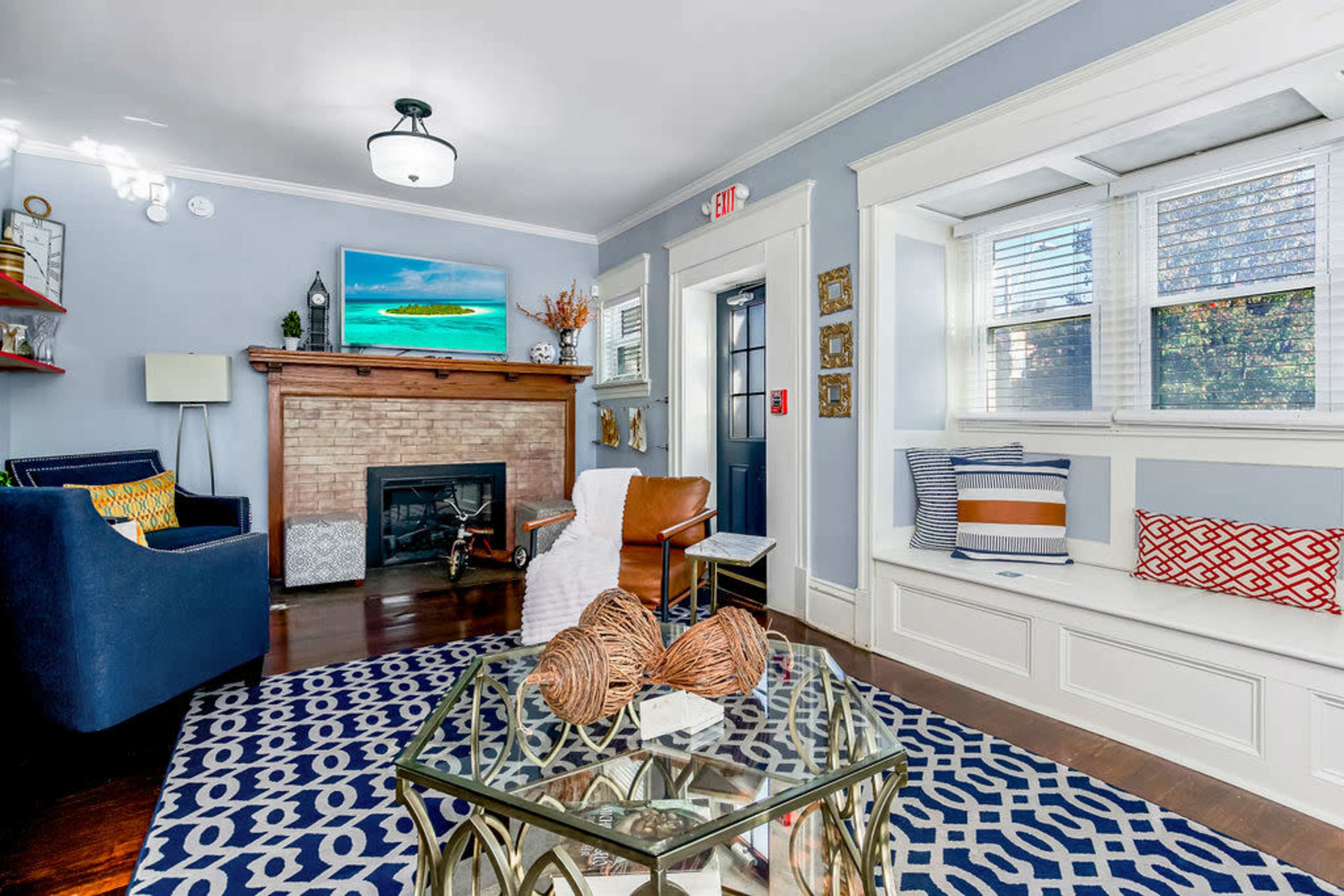 The image shows a cozy living room featuring a fireplace, a television mounted on the wall, a patterned area rug, and a mix of seating options, including a blue armchair and a leather chair.