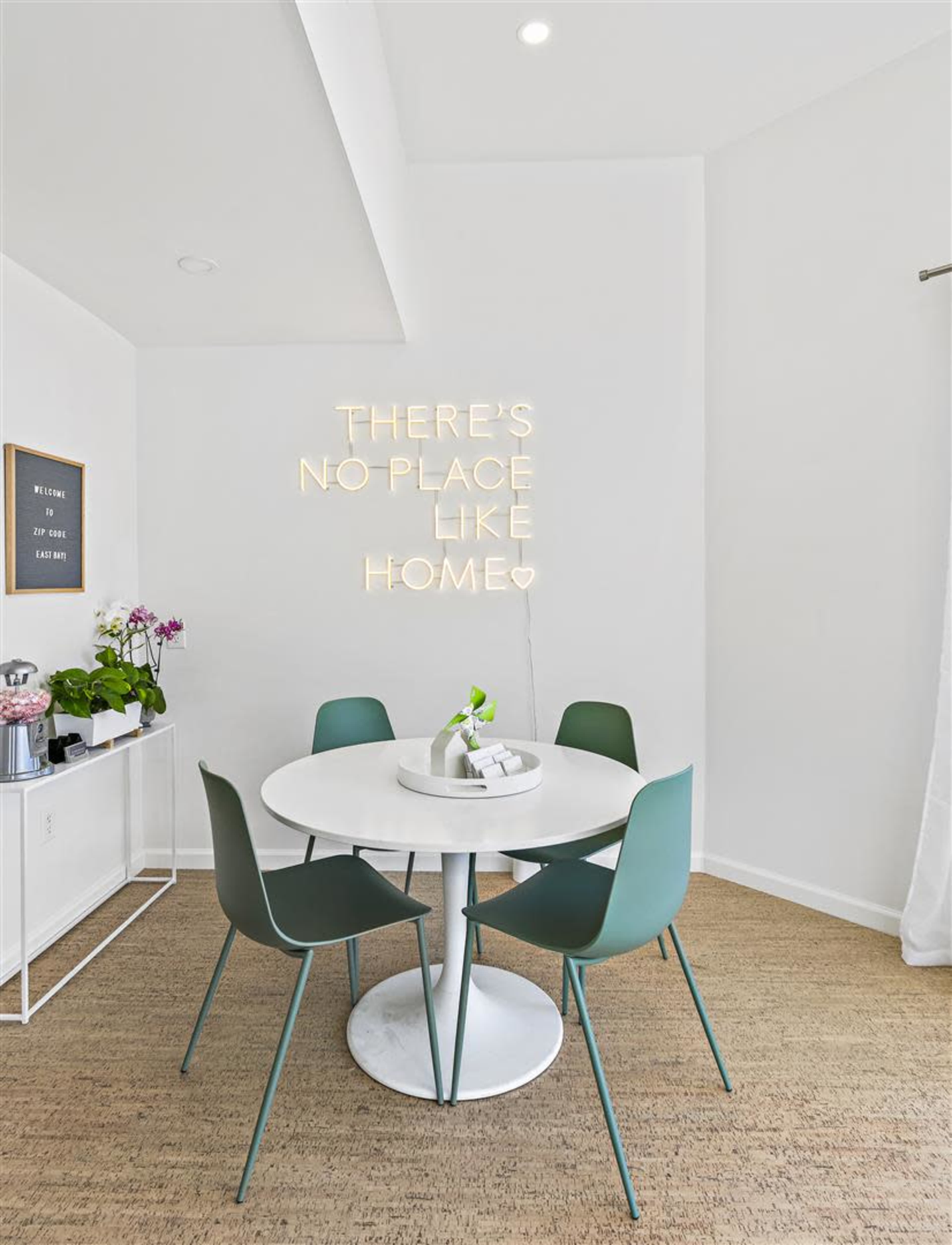 A round white dining table with four green chairs is set against a white wall featuring illuminated text that reads, "THERE'S NO PLACE LIKE HOME."