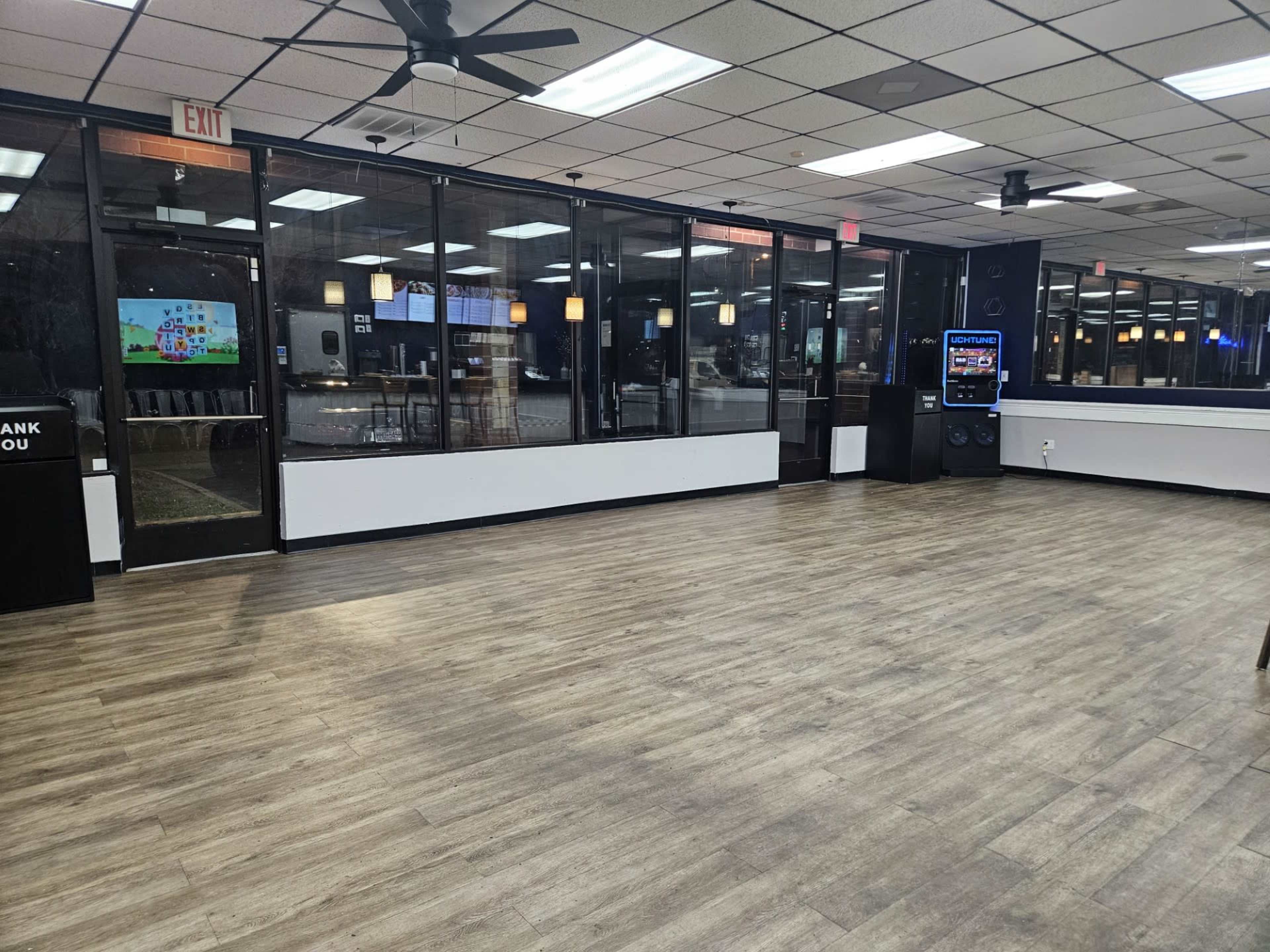 The image shows a spacious indoor area with wooden flooring, large windows, and various seating arrangements that are currently unoccupied.