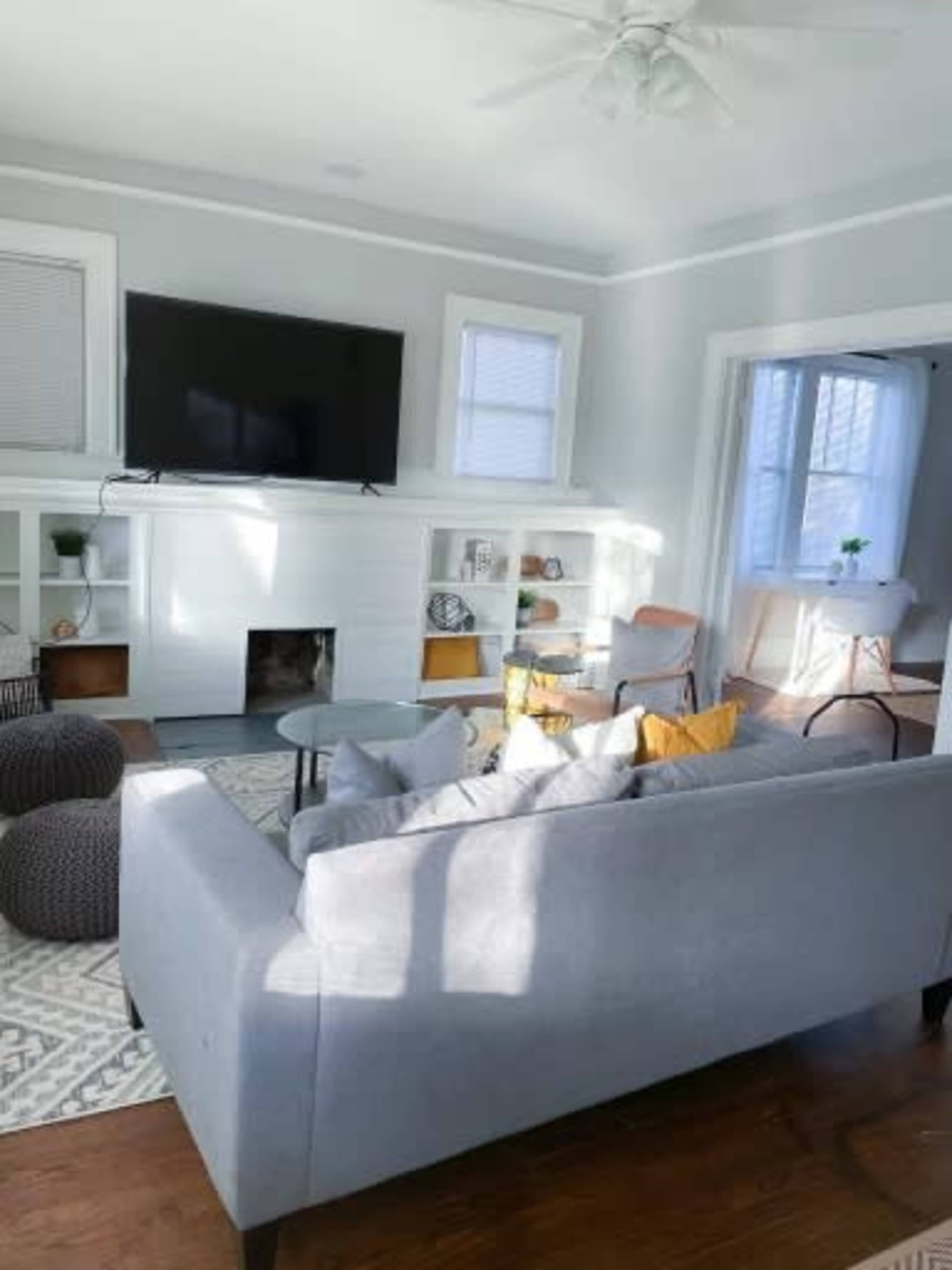 A modern living room features a gray sofa, a circular coffee table, and a large TV mounted on a white wall, with built-in shelves and a window letting in natural light.