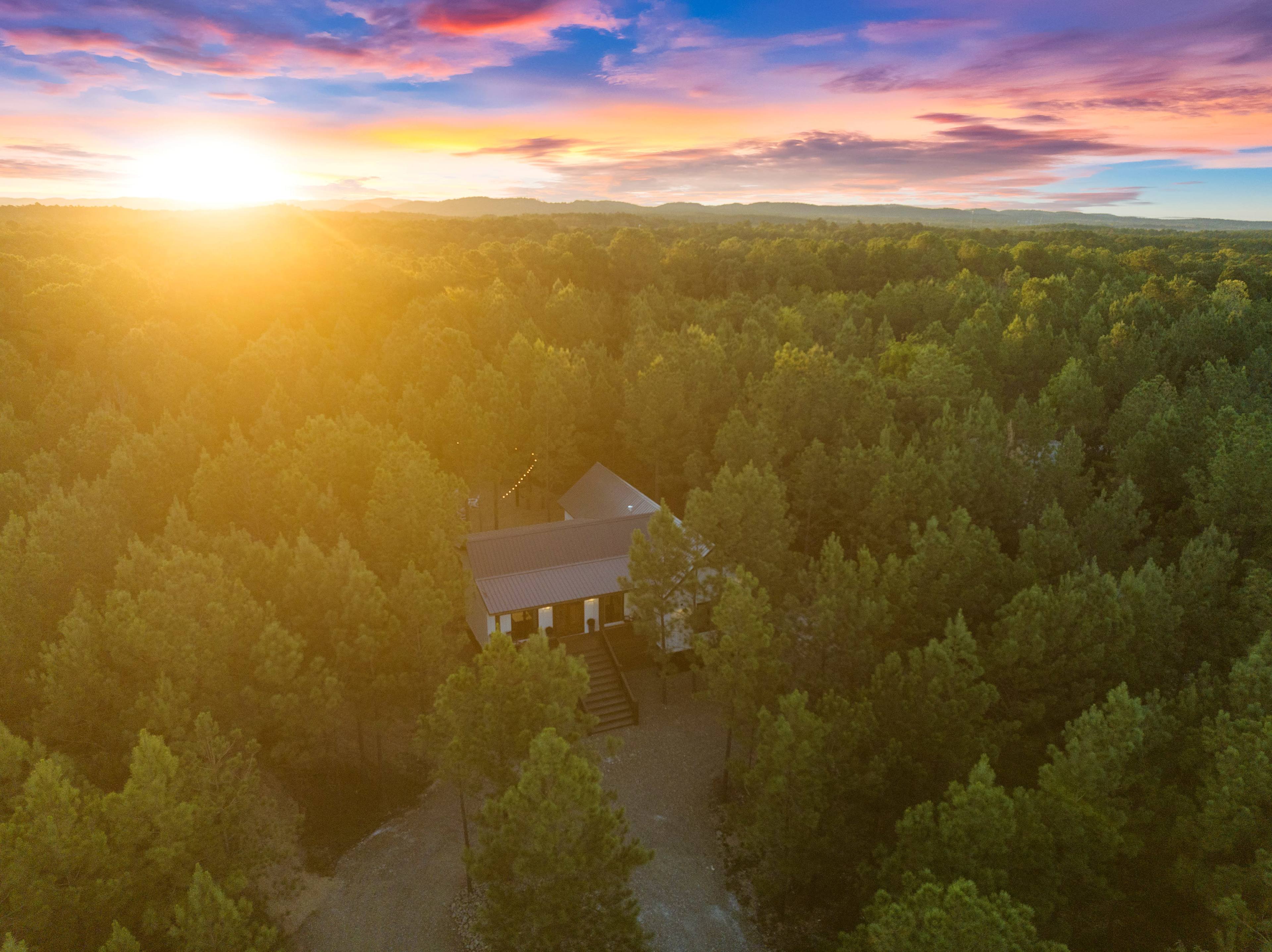 A house is situated among dense pine trees, illuminated by a sunrise in the background.