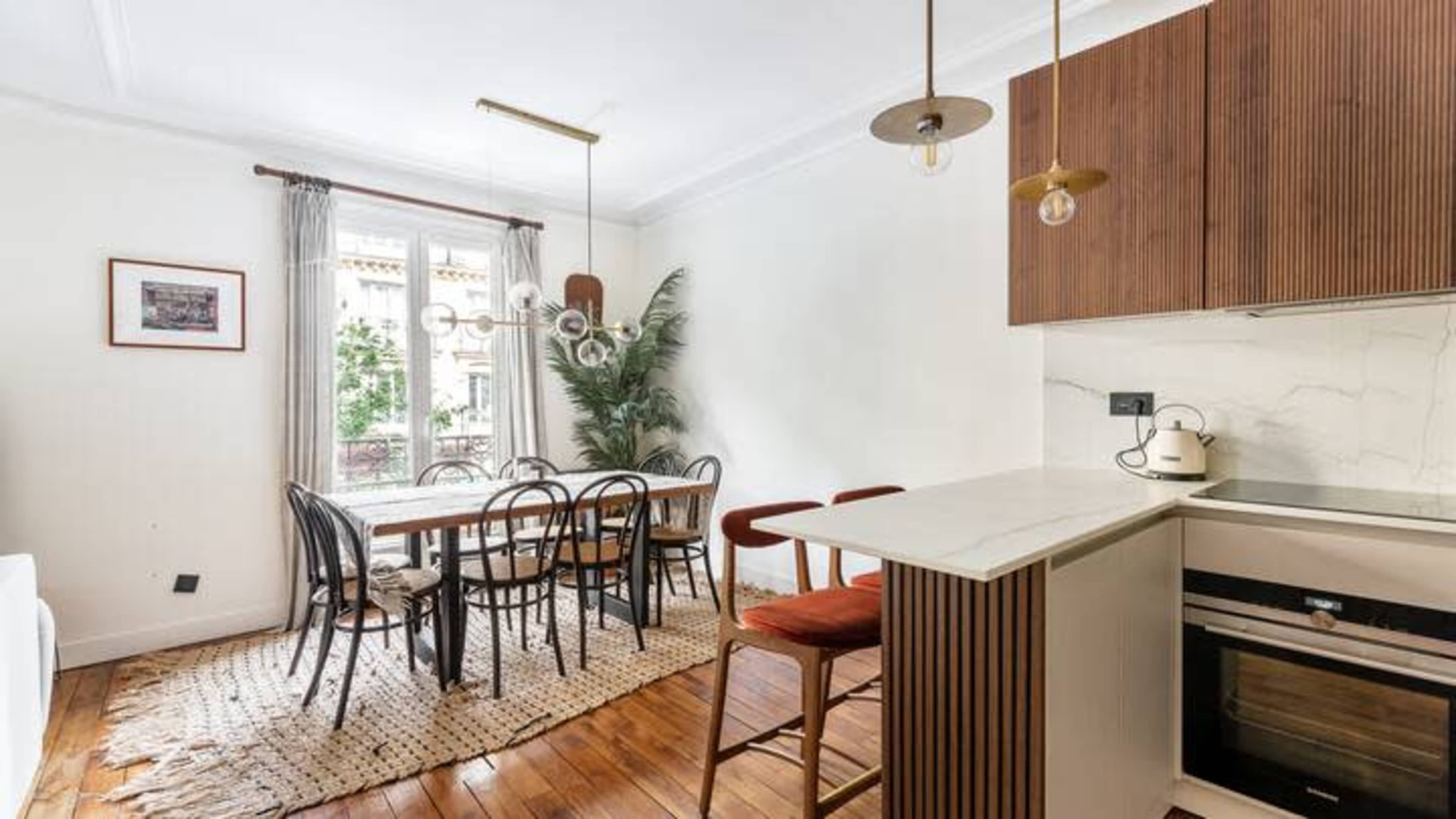 A modern dining area features a wooden table surrounded by black chairs, with a kitchen bar and large windows letting in natural light.