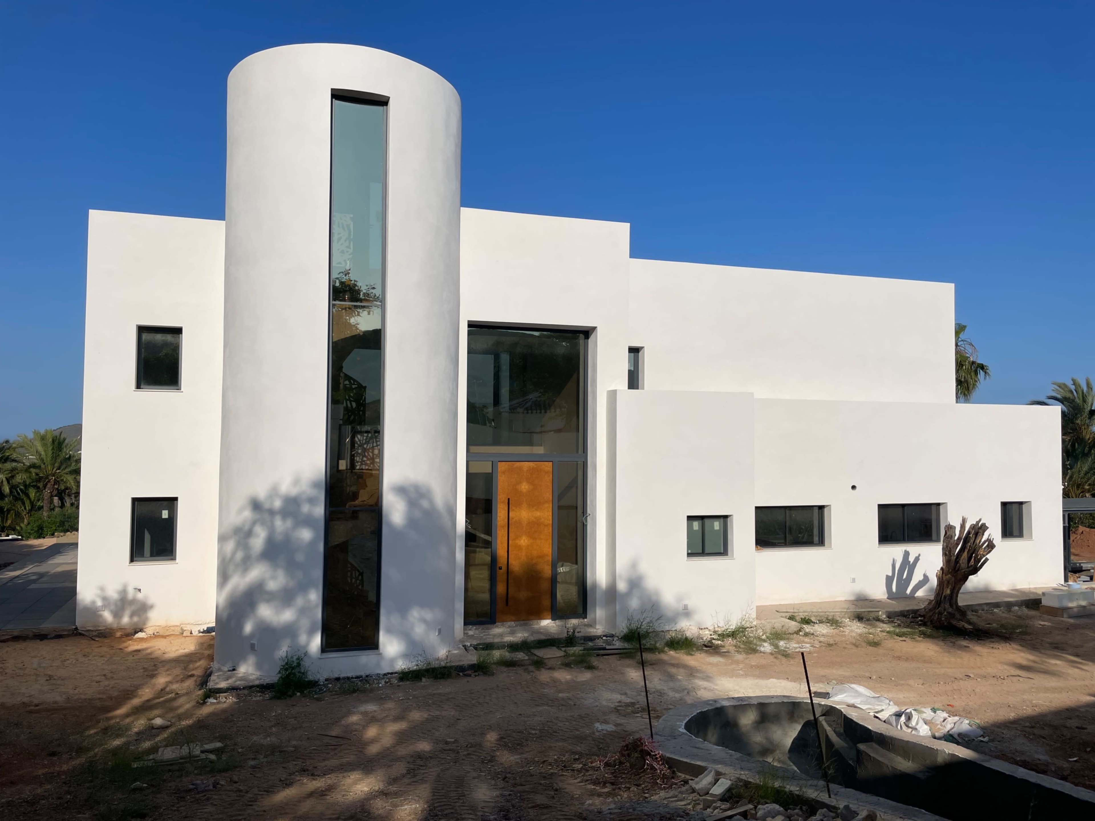 A modern white house features a cylindrical structure, large glass panels, and an unfinished area in front.