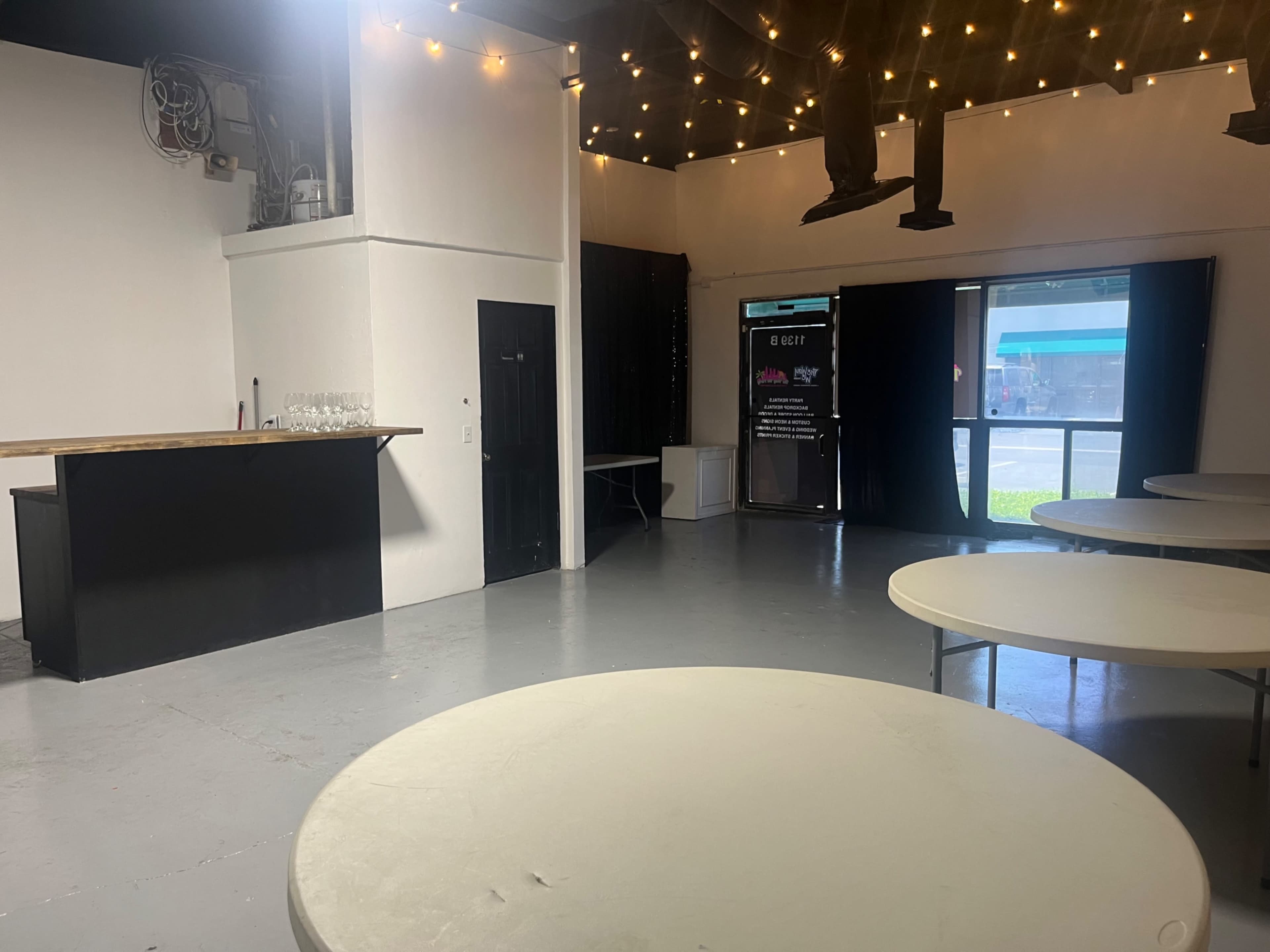 The image shows an empty indoor space featuring round tables, a bar area against one wall, and large windows revealing an exterior view.