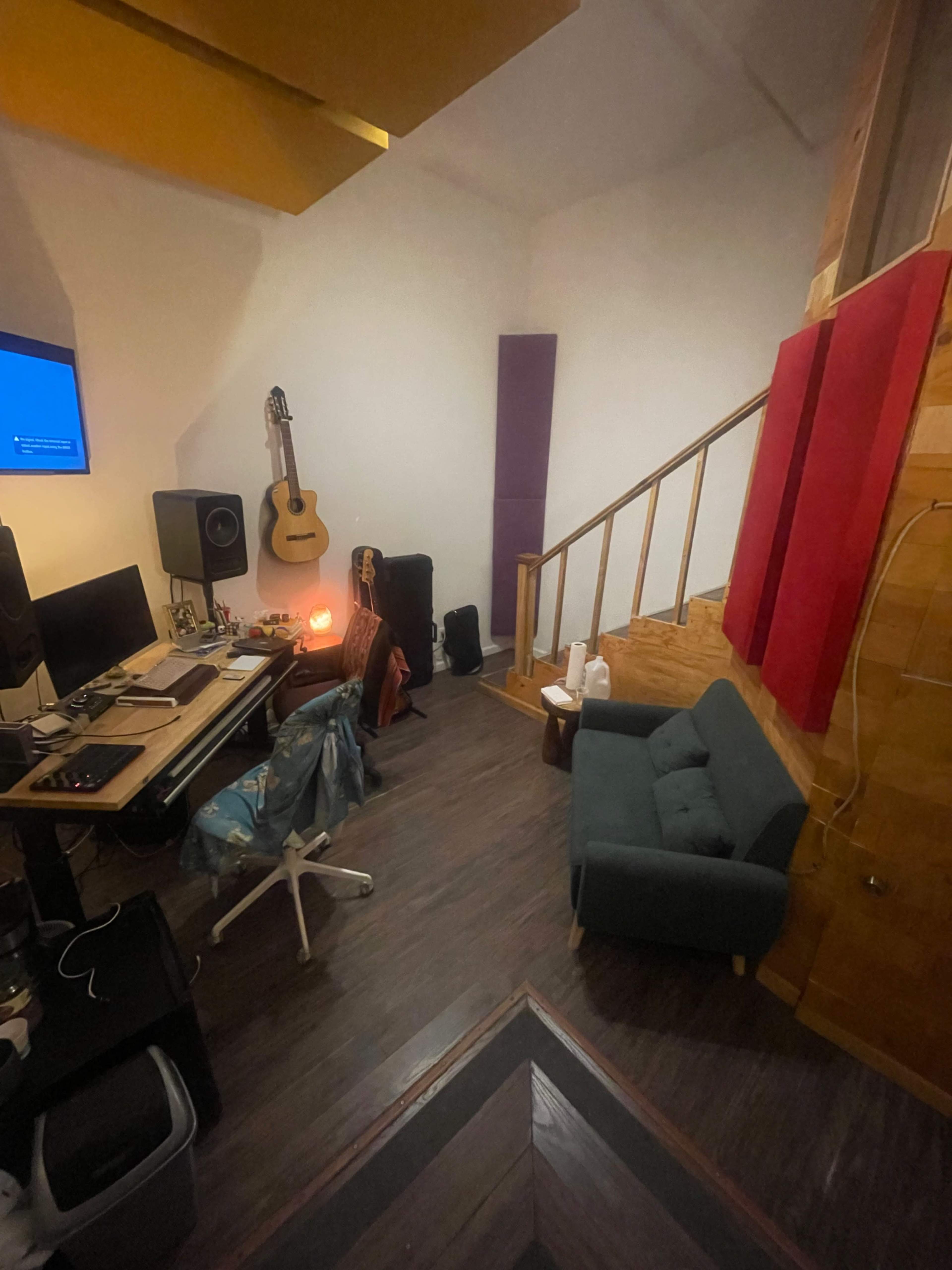 The image shows a small music studio or workspace with a computer setup, a guitar, a sofa, and decorations on the walls.