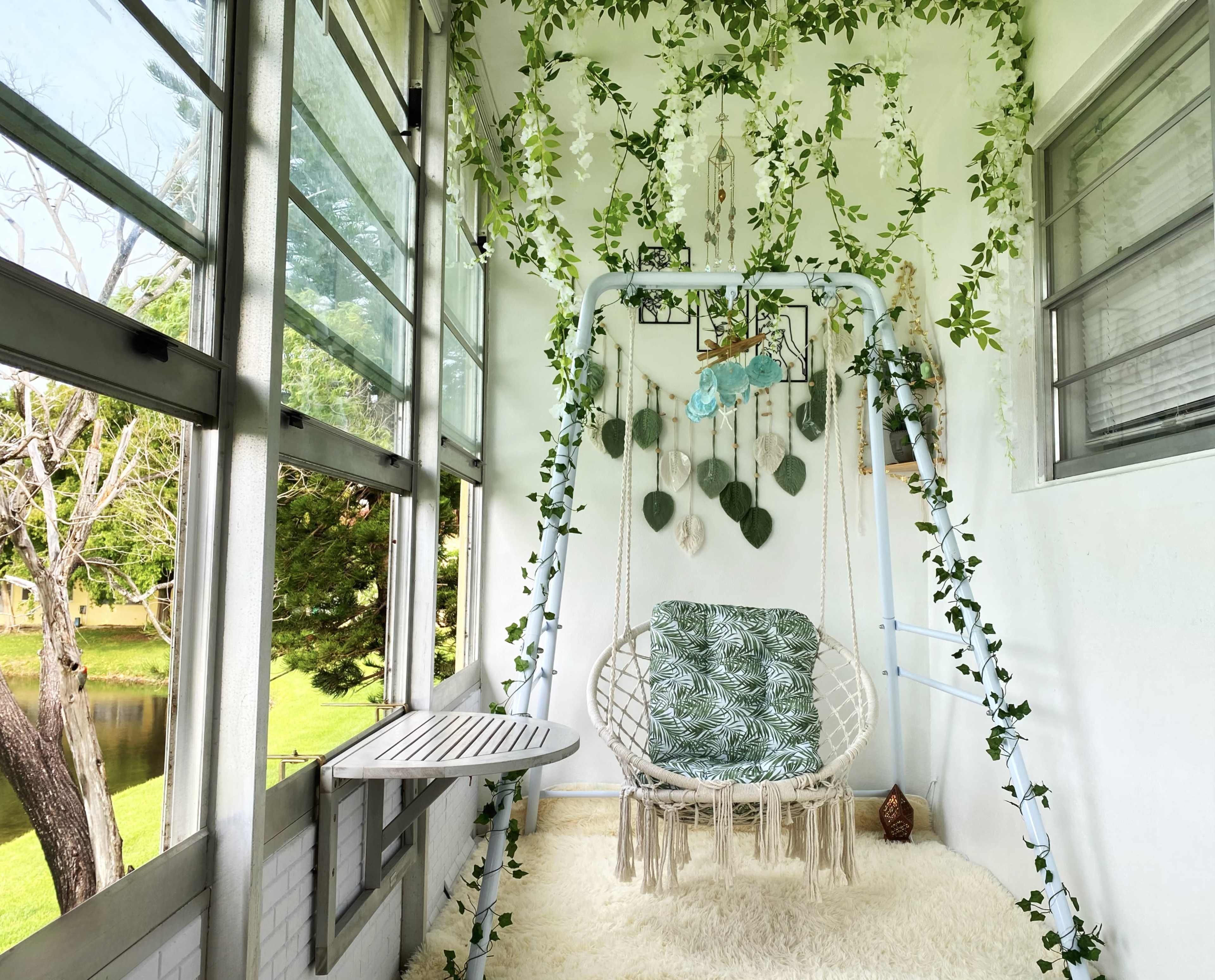 A swing chair is positioned on a sunlit balcony adorned with greenery and soft decor overlooking a serene outdoor view.