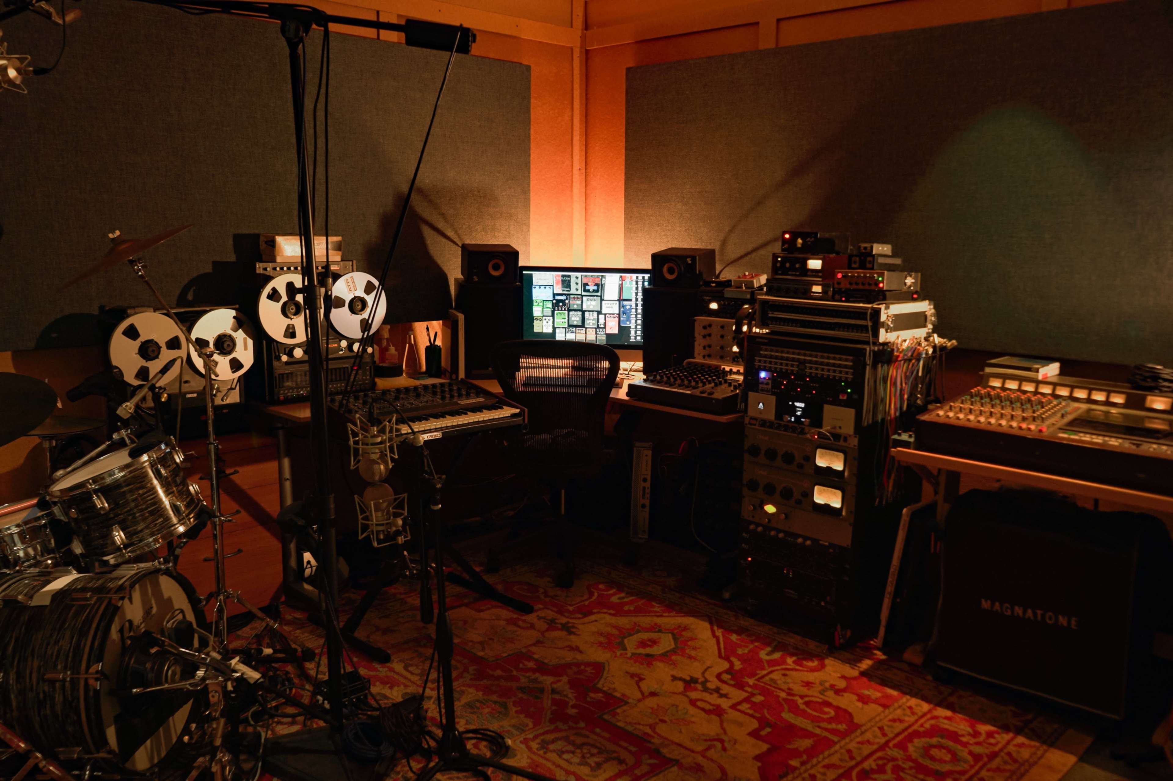 The image shows a music studio with recording equipment, including a drum set, reel-to-reel tape machines, and a computer setup in a soundproofed room.