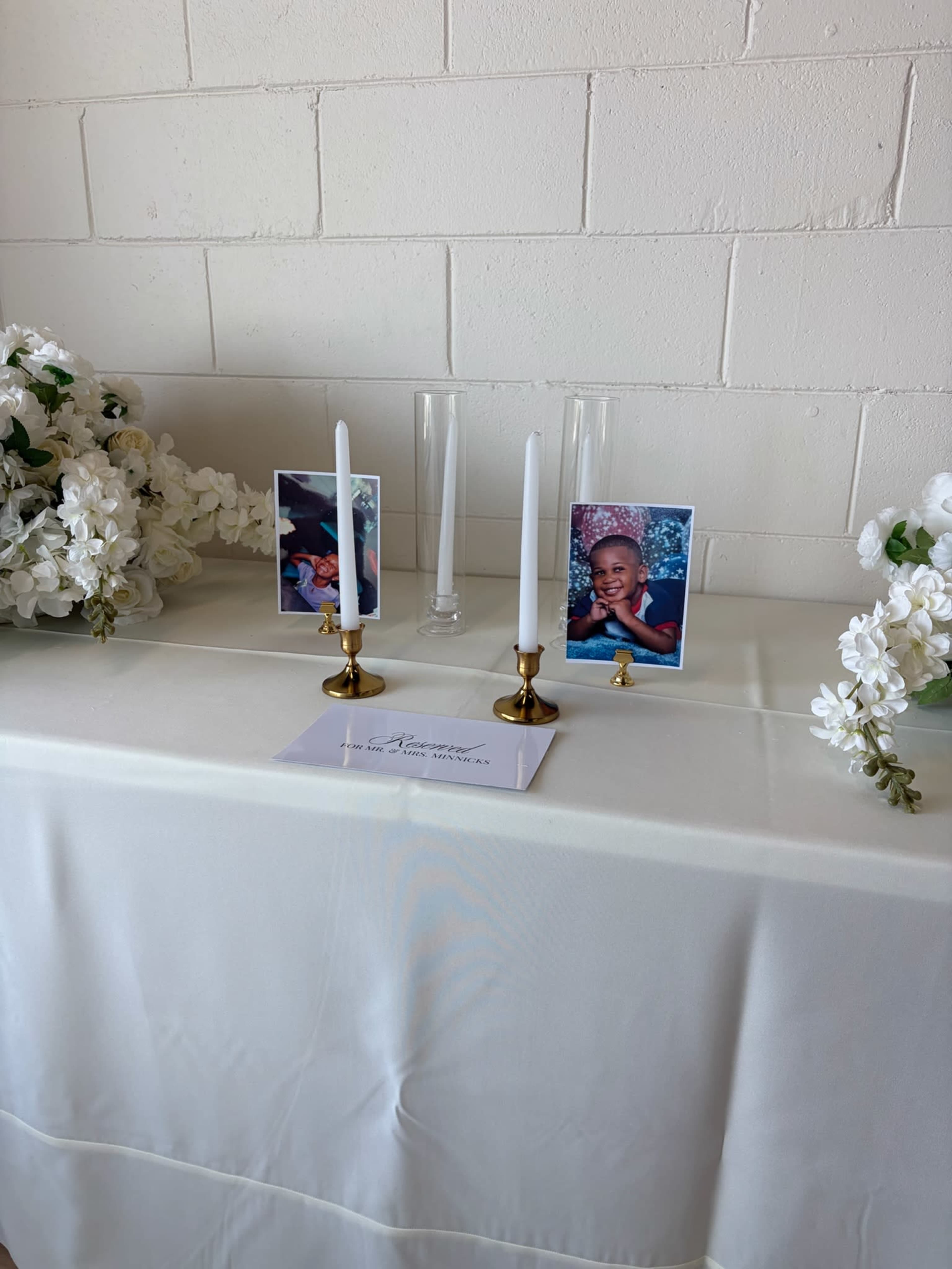 The image shows a table covered with a white cloth, adorned with two candles, two framed photographs of a child, a clear vase, and artificial white flowers on either side.