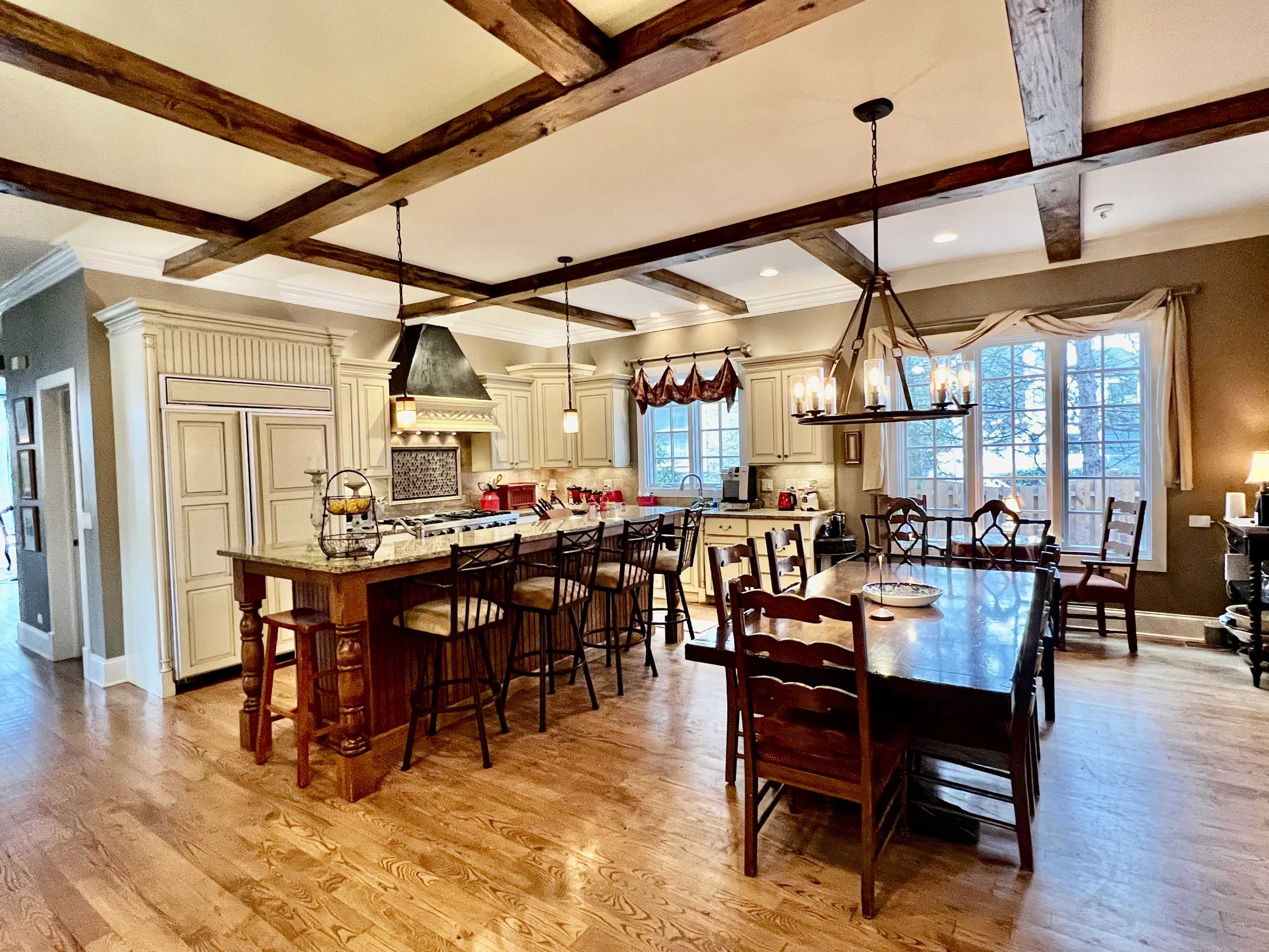 The image shows a spacious kitchen with a large island, wooden beams on the ceiling, and a dining area with a long wooden table and chairs.