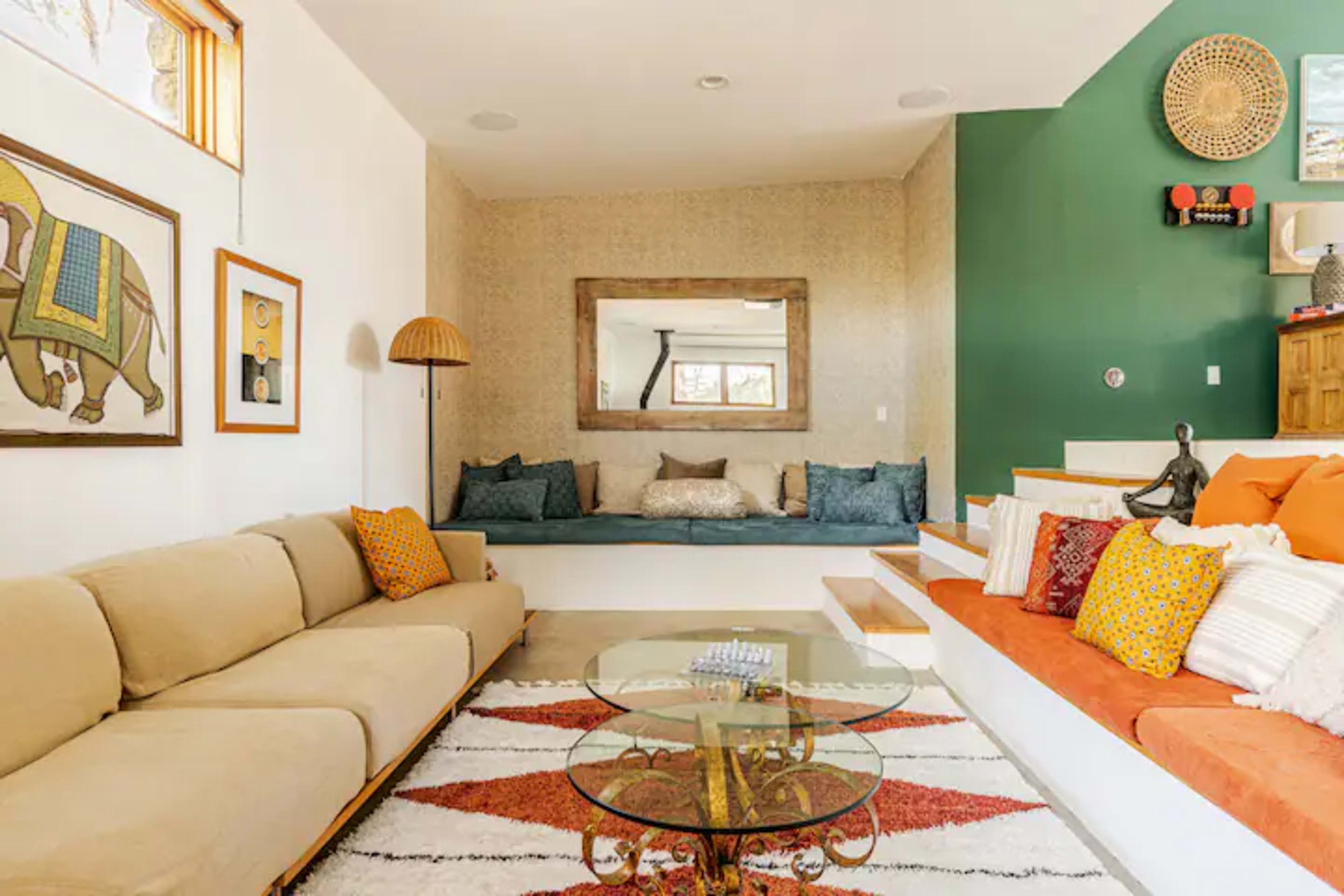 The image shows a modern living room featuring a beige sofa, a glass coffee table, a colorful area rug, and a seating area with cushions against the wall, complemented by decorative wall art and a mirror.