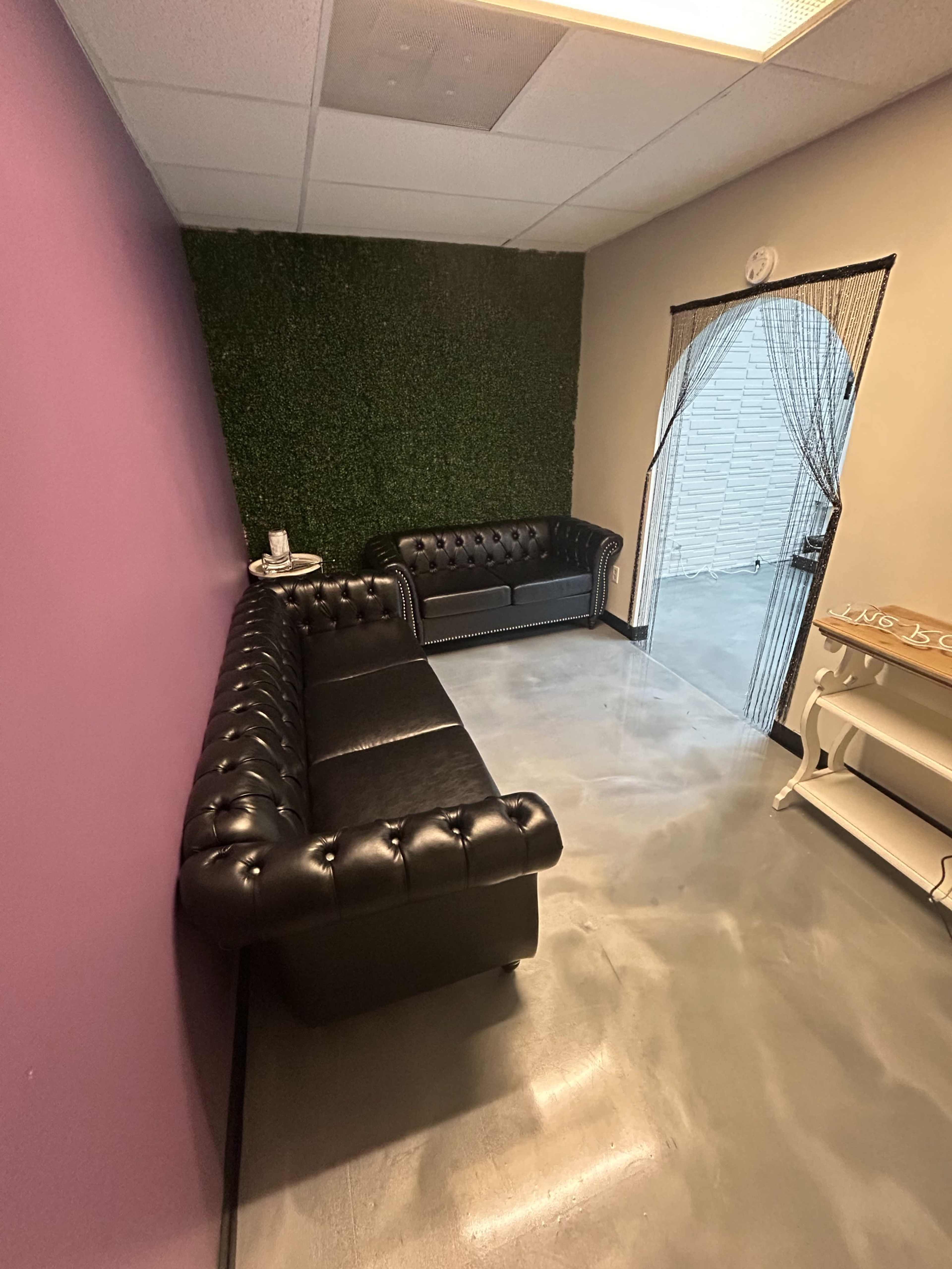 A small waiting area with a black leather sectional sofa against a purple wall and a door leading to another room, complemented by a green wall covering.