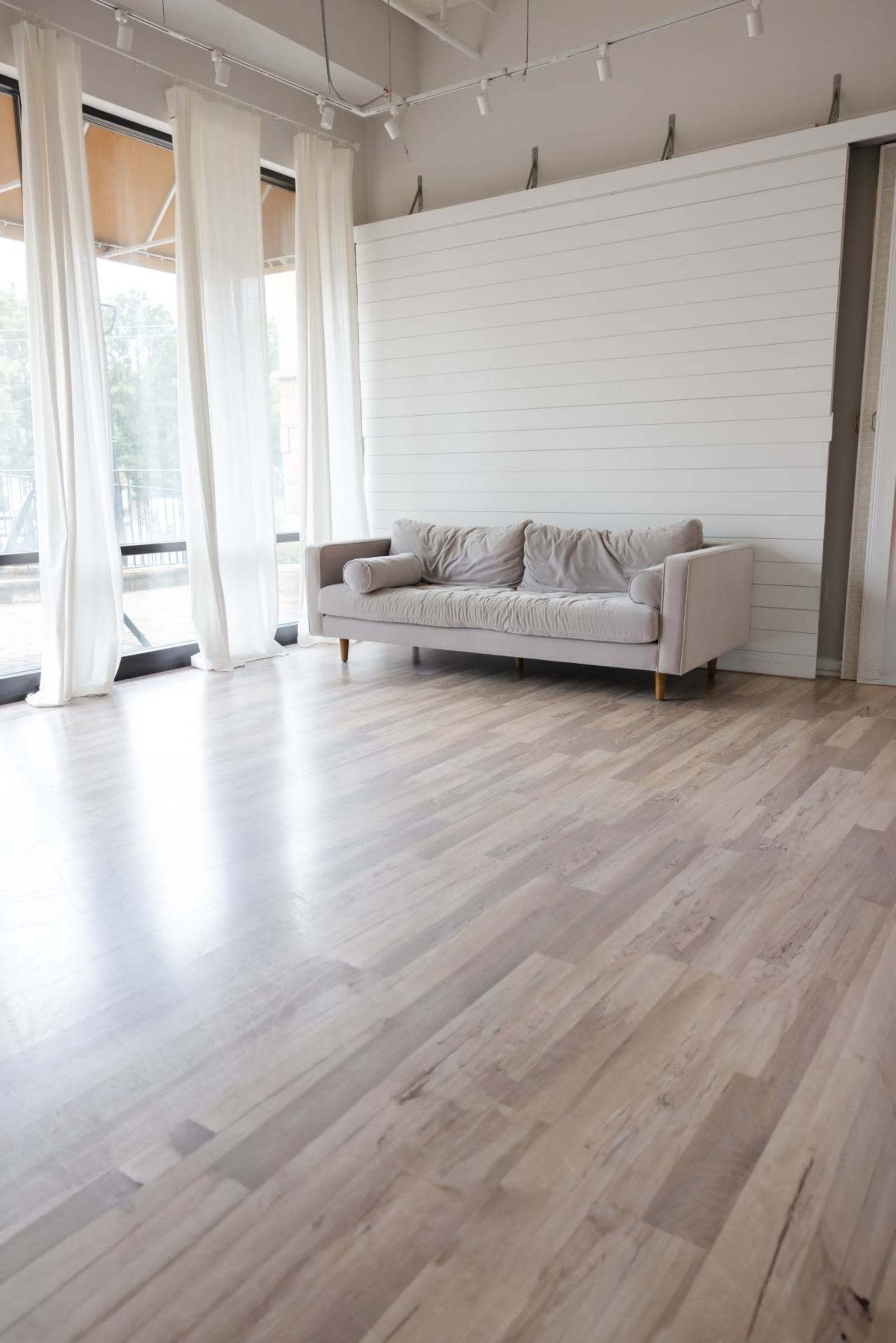 The image shows a light-colored wooden floor with a soft gray couch against a white wooden wall, surrounded by sheer curtains in a bright, open space.