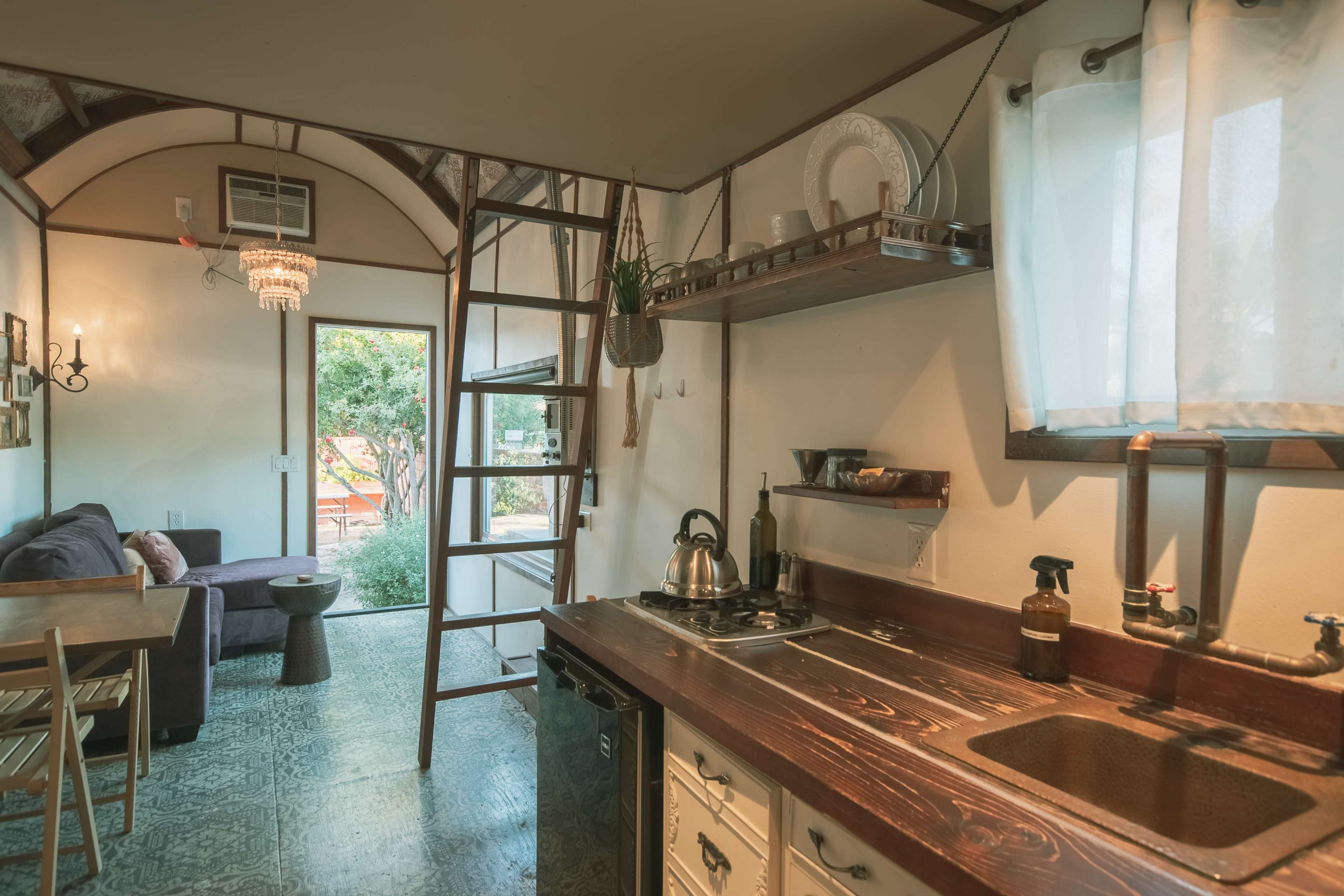 The image shows a compact kitchen area with wooden cabinetry, a small dining table, and a ladder leading to an upper level, next to a doorway opening to an outdoor space.