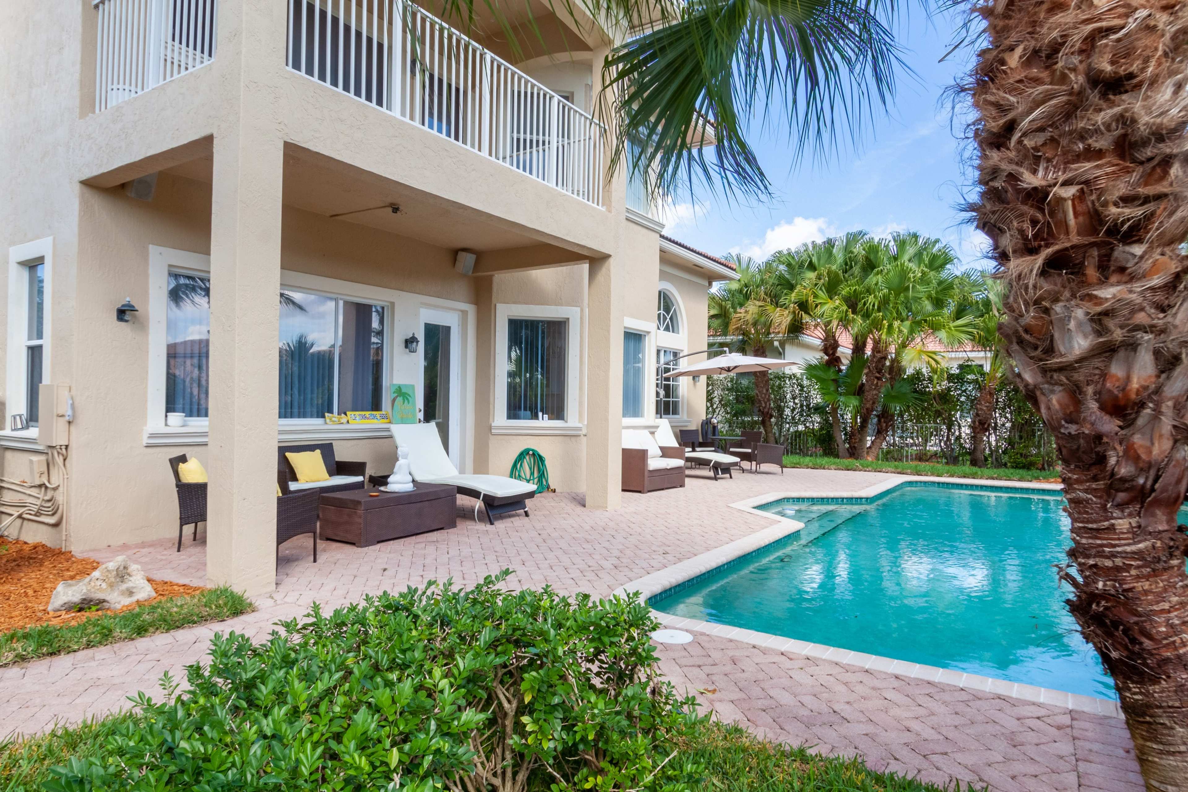 A two-story house with a swimming pool and lounge chairs sits in a landscaped yard surrounded by palm trees.