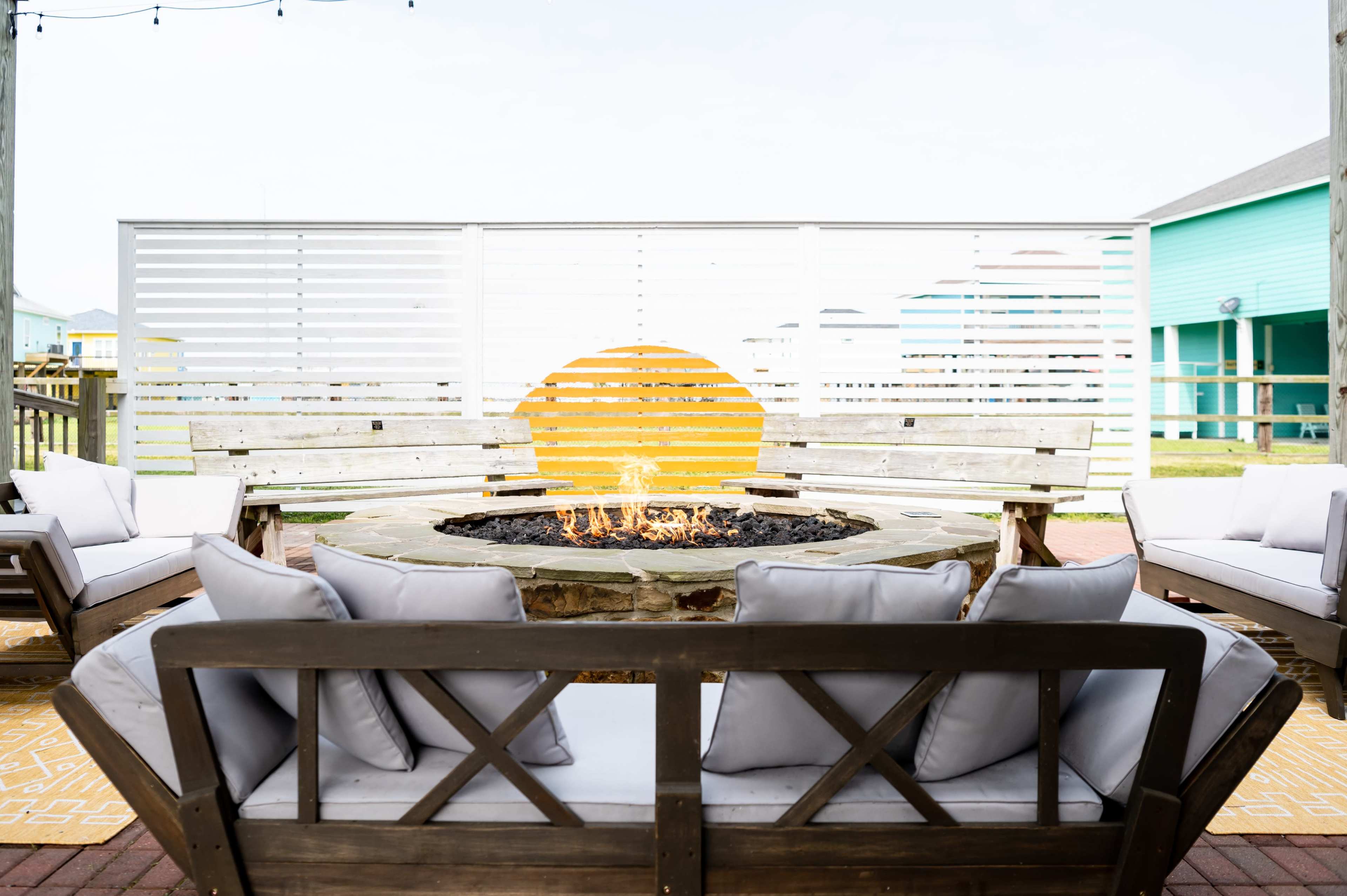 A fire pit surrounded by wooden benches and cushions is set against a backdrop of a white slatted wall with a yellow circular design, indicating sunset.
