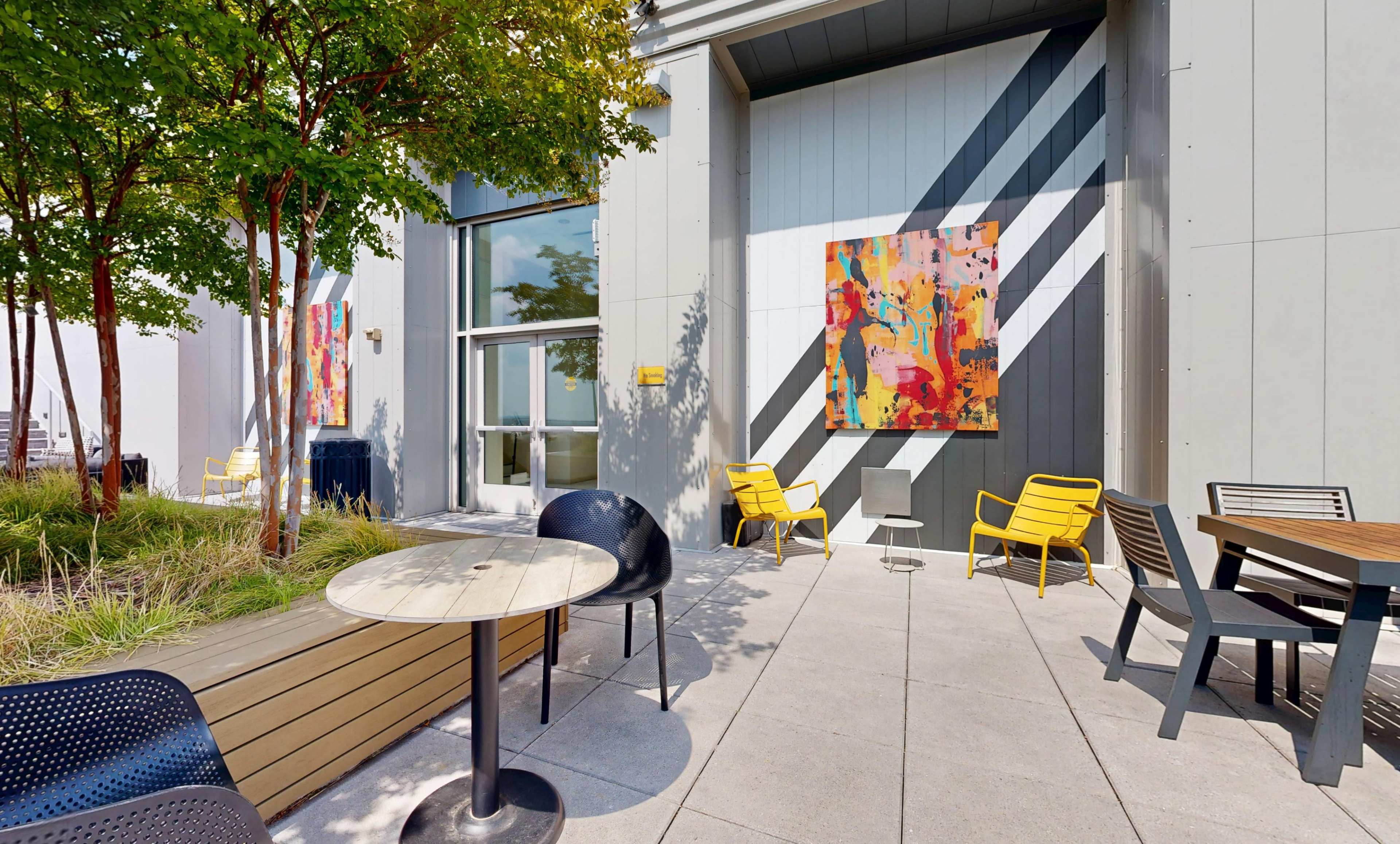 The image shows a modern outdoor seating area with colorful artwork on the walls, yellow chairs, and various tables surrounded by greenery.