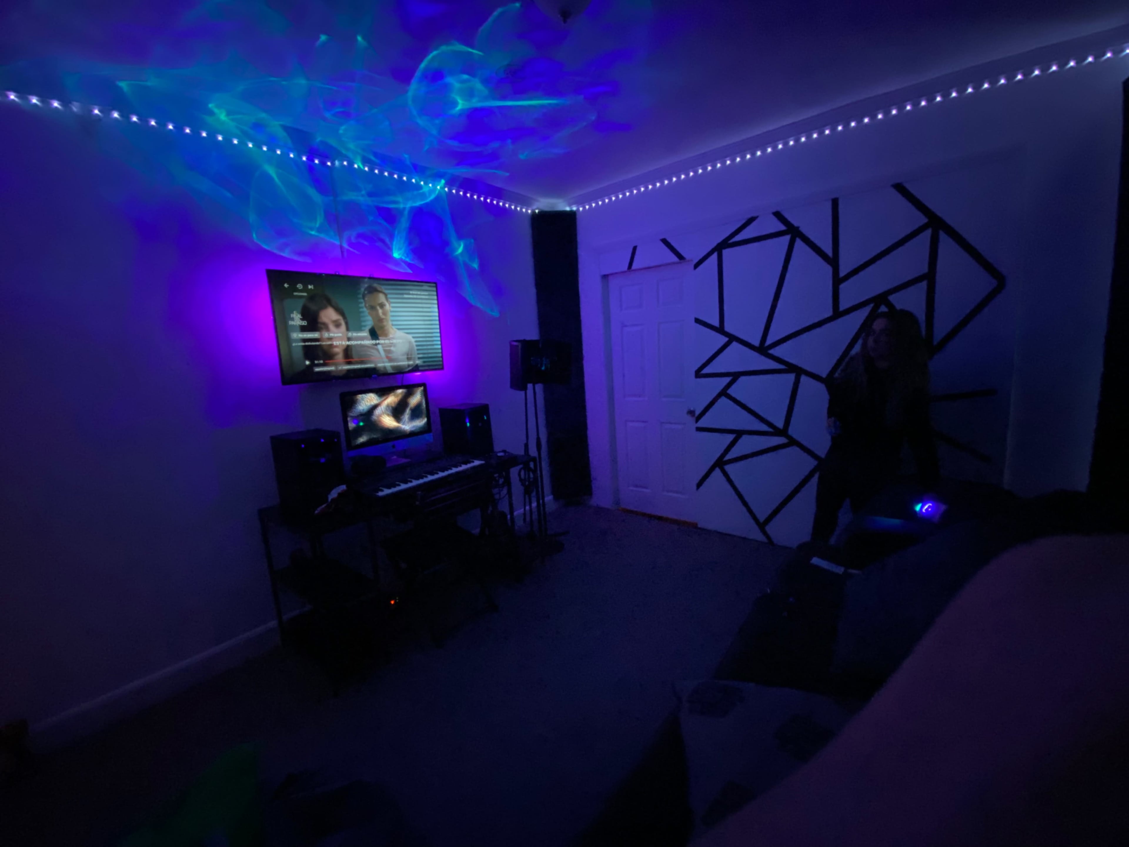 A dimly lit room with colorful lighting effects, featuring a television, a computer setup, and a person standing near a decorated wall.