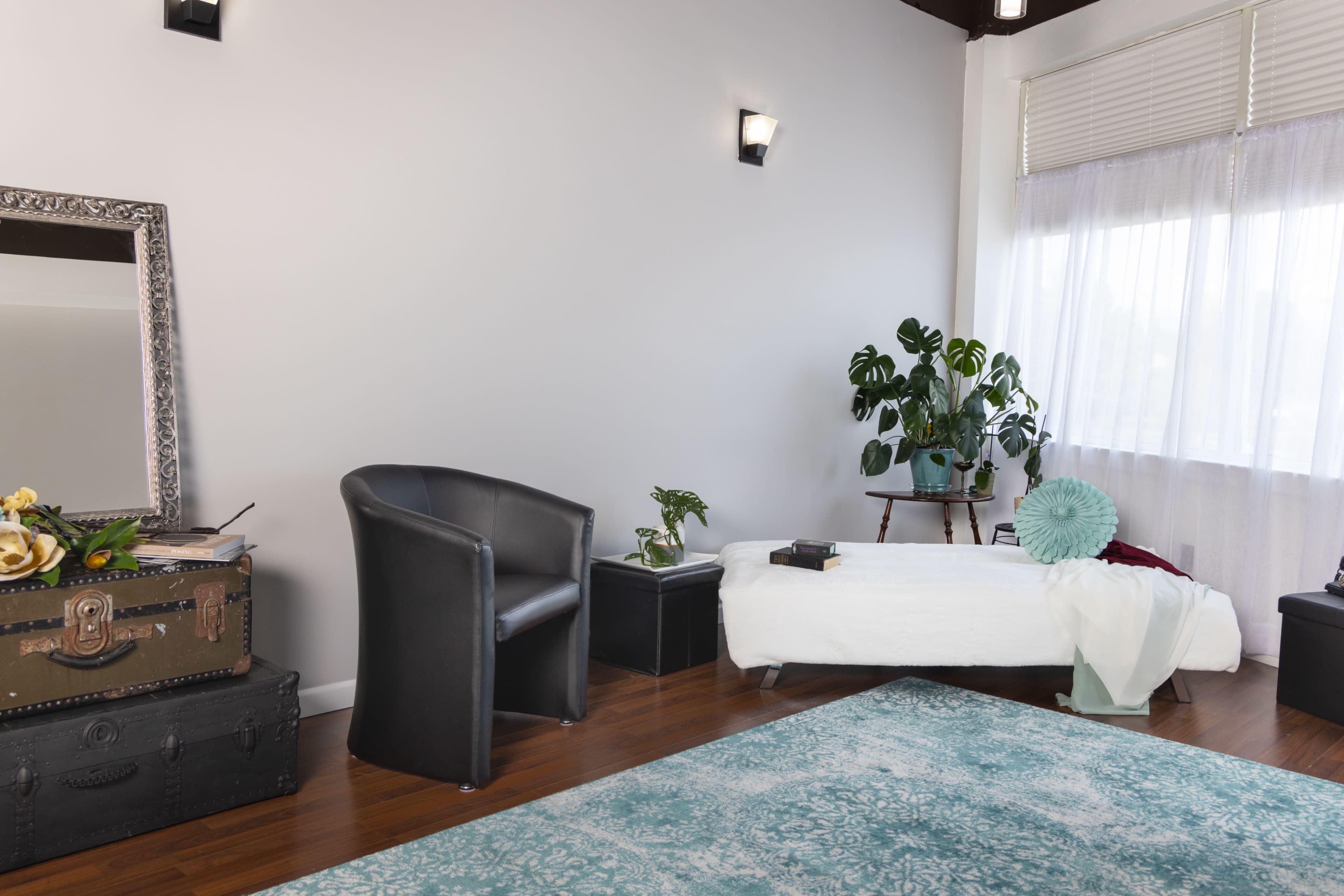 The image shows a minimalist room with a white daybed, a black chair, a large mirror, and a window covered with sheer curtains, accompanied by potted plants and decorative items.