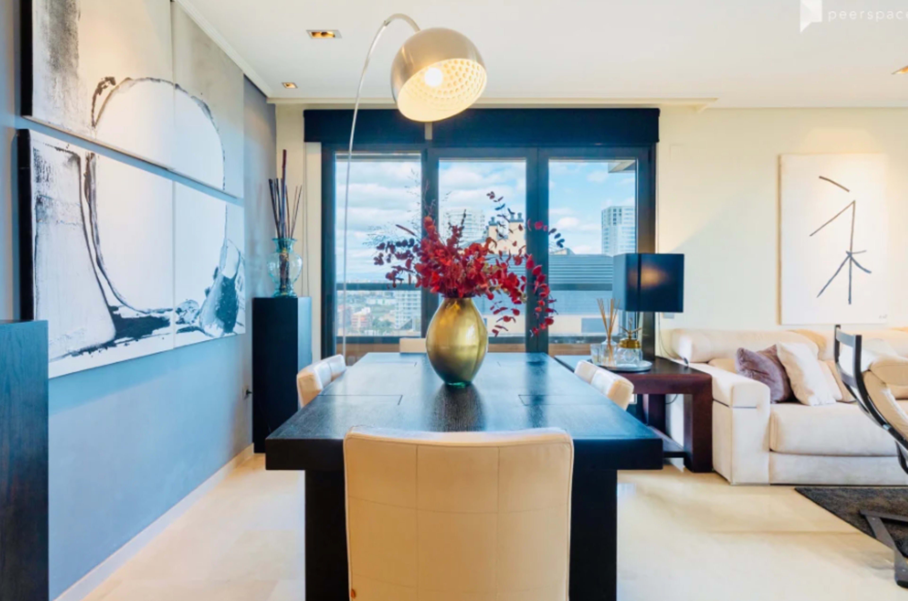 The image shows a modern interior featuring a dining table with a vase of red flowers, large windows with city views, and contemporary artwork on the walls.