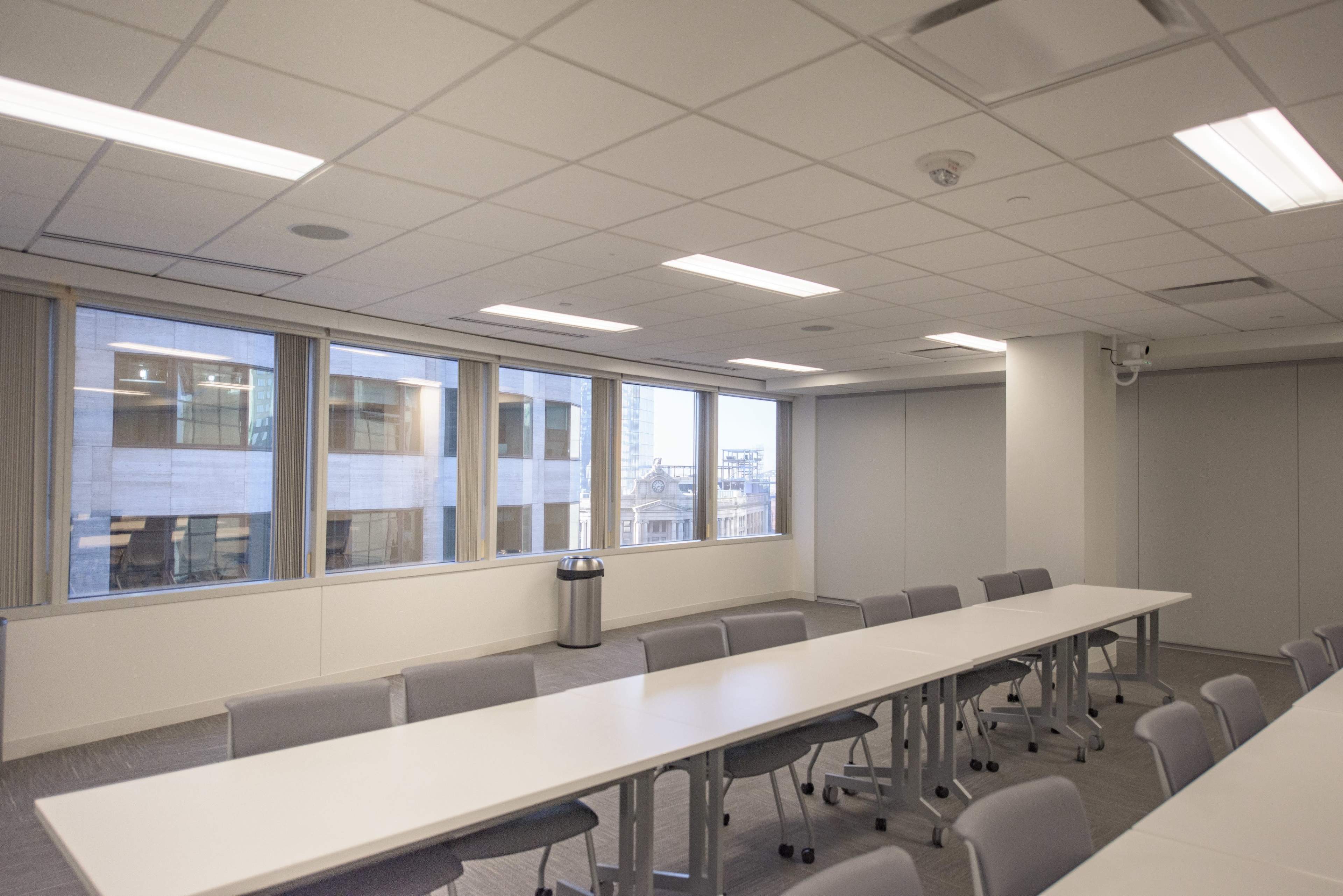A conference room features a long white table surrounded by gray chairs, with large windows providing a view of the outside.