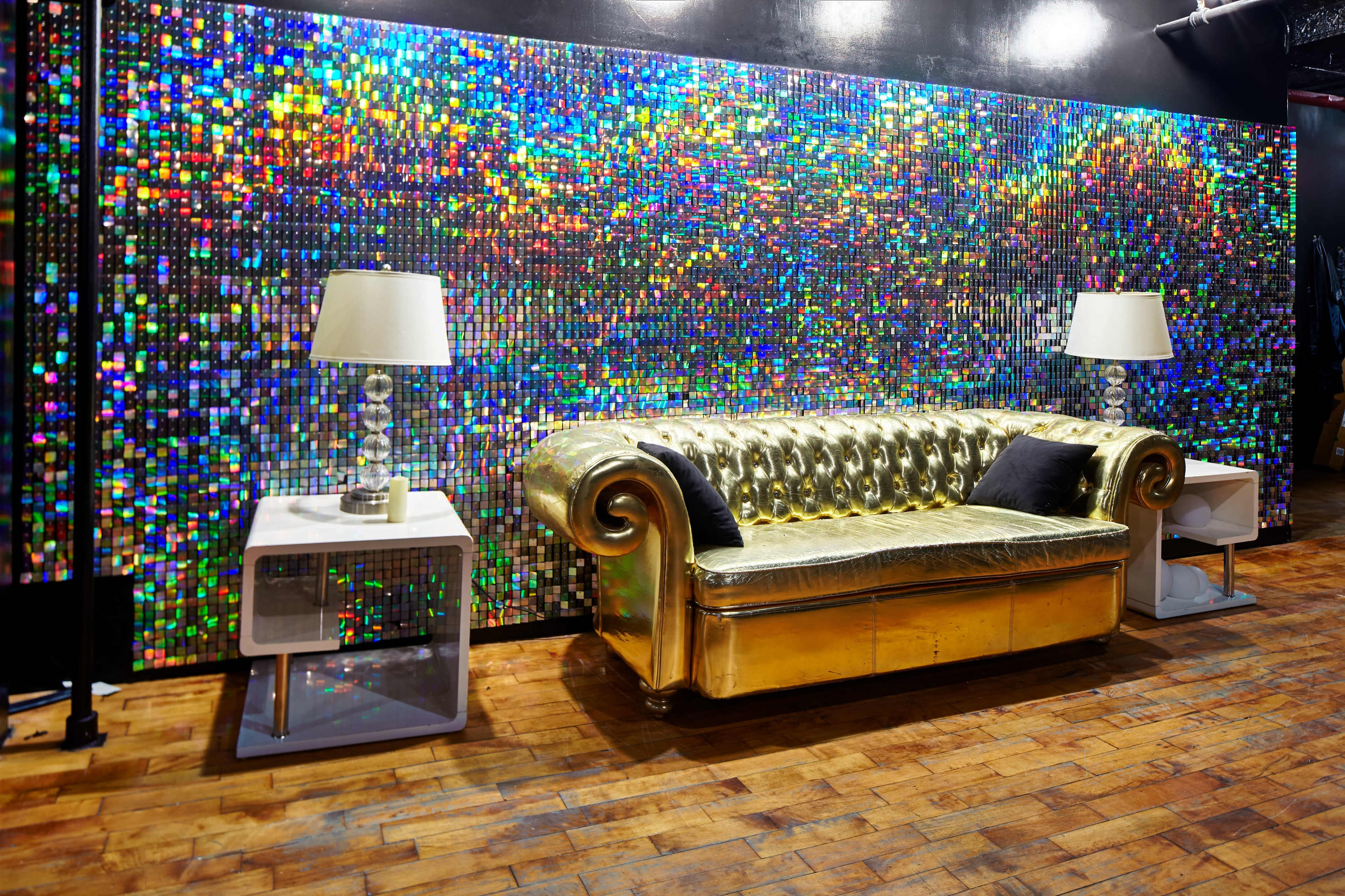 A gold couch with decorative cushions is positioned in front of a colorful, shimmering wall, with two white side tables and lamps on either side.