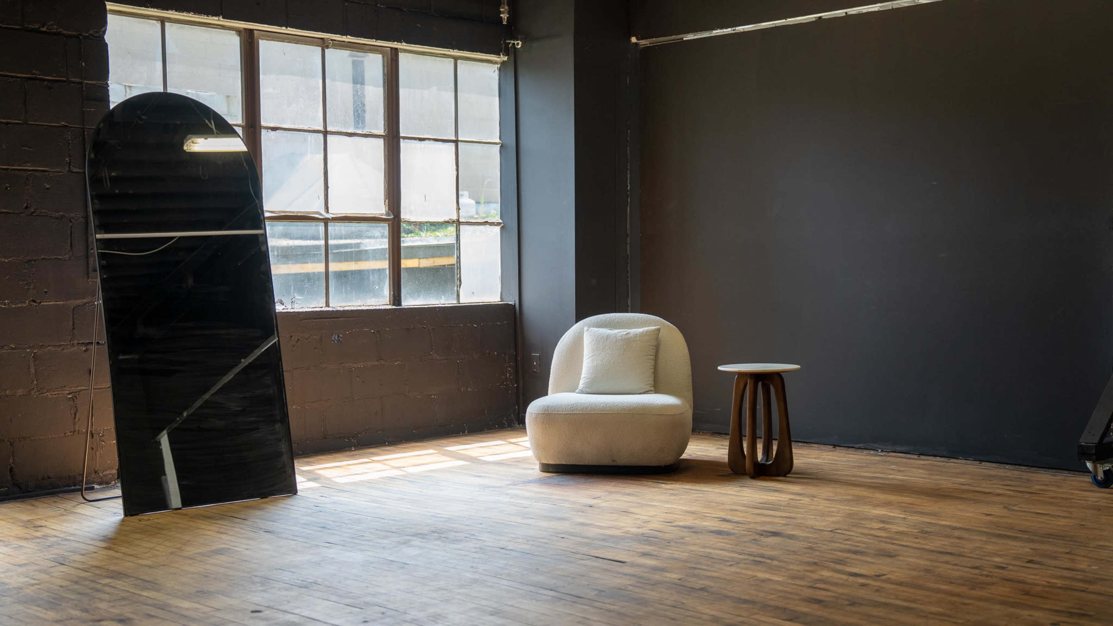 The image shows a modern, minimalist space featuring a curved chair, a small wooden side table, and a large, reflective mirror next to a window.
