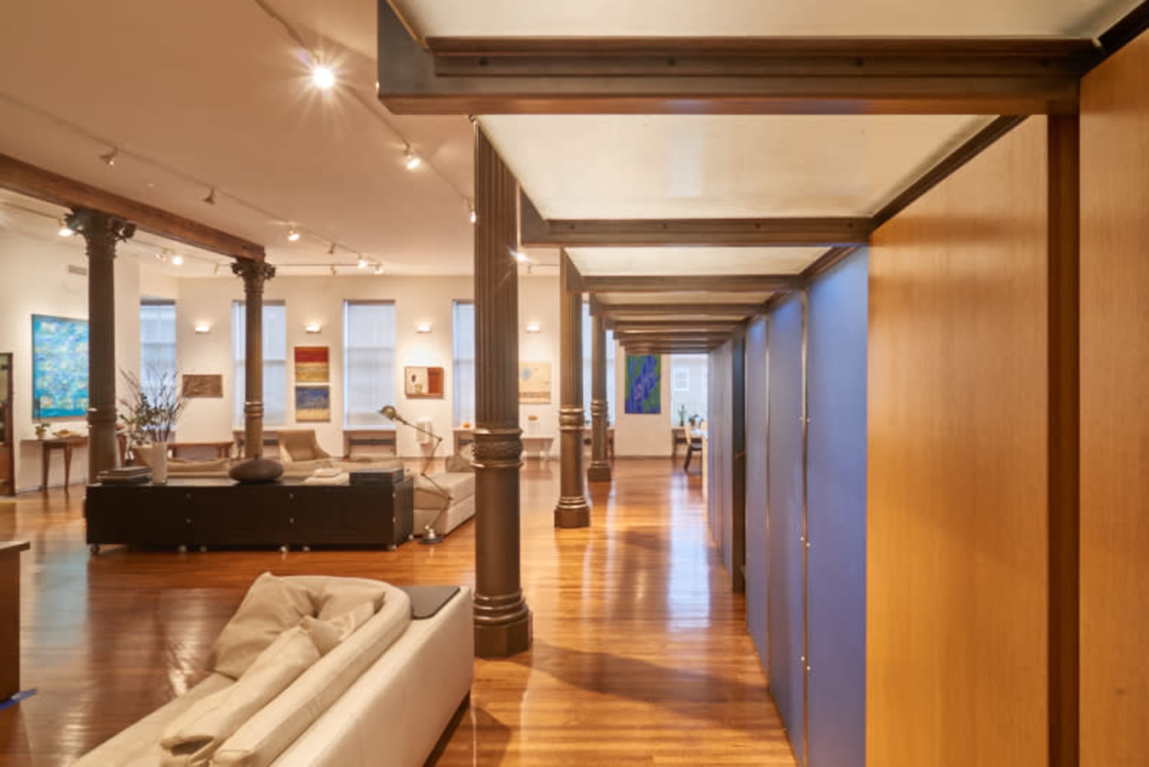 A spacious interior of a modern art gallery with wooden floors, columns, and various artwork displayed on the walls.