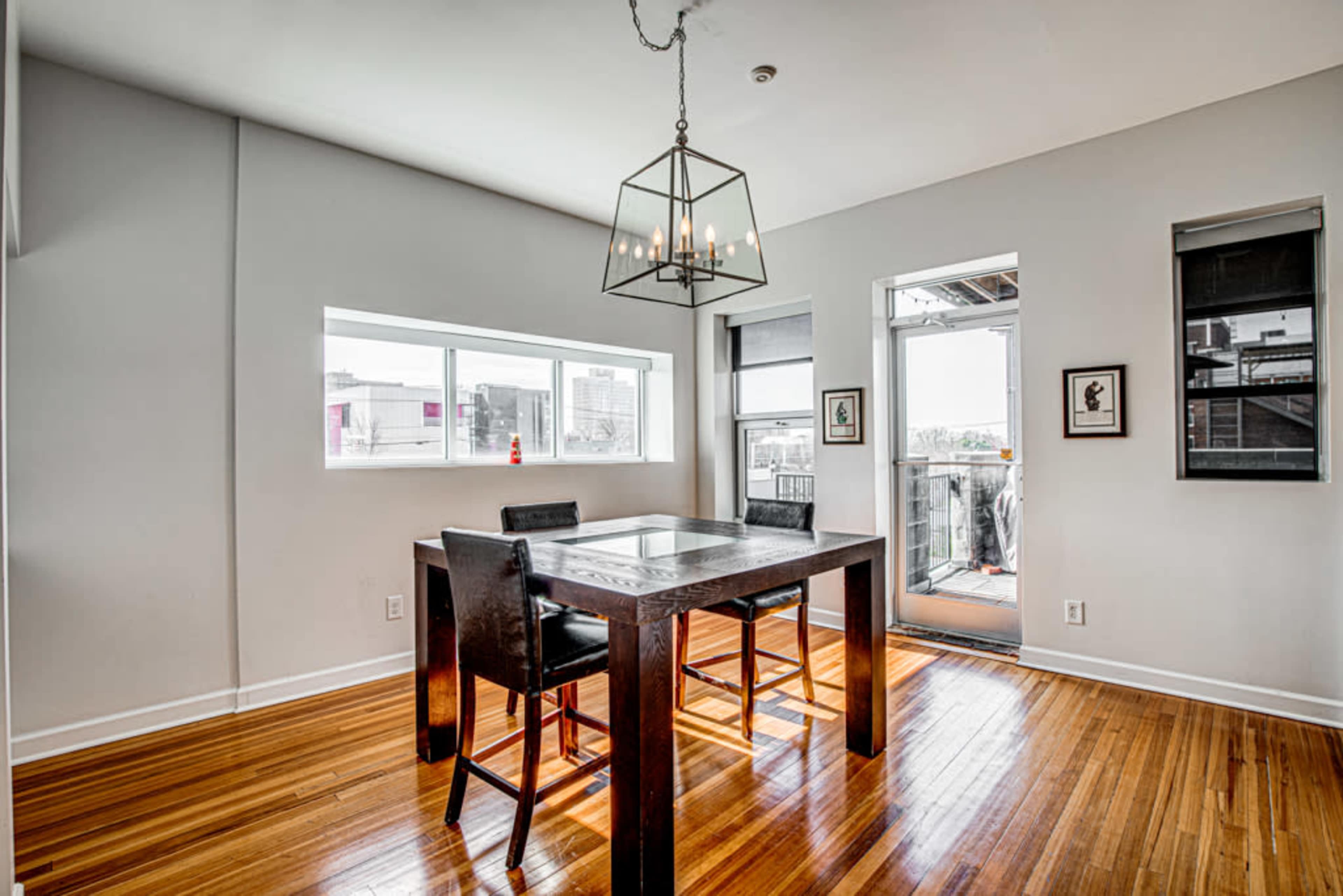 A dining area features a square table with four chairs and a large pendant light, illuminated by natural light from two windows.