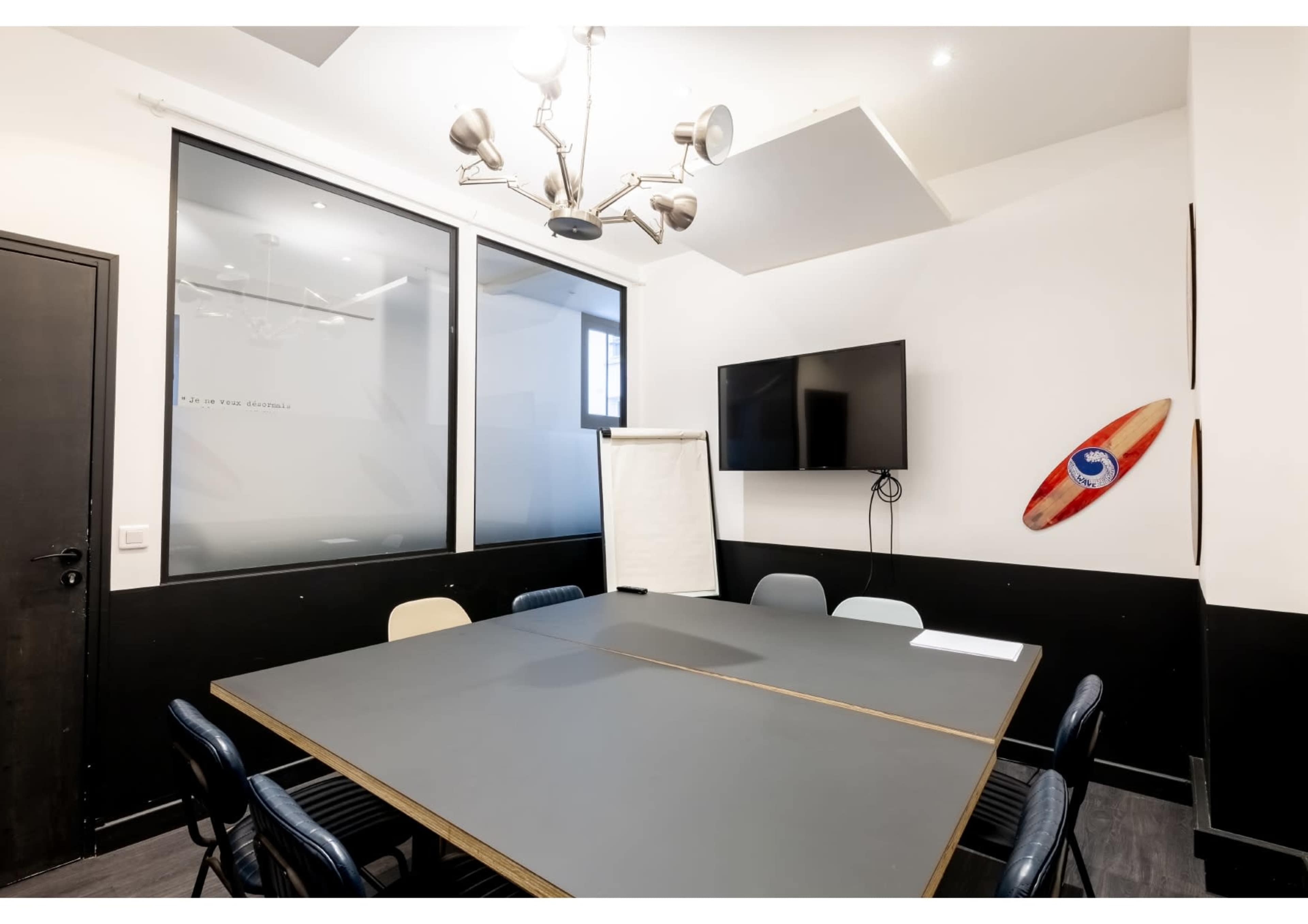A modern meeting room features a large gray table surrounded by chairs, a flat-screen TV on the wall, and a whiteboard stand.