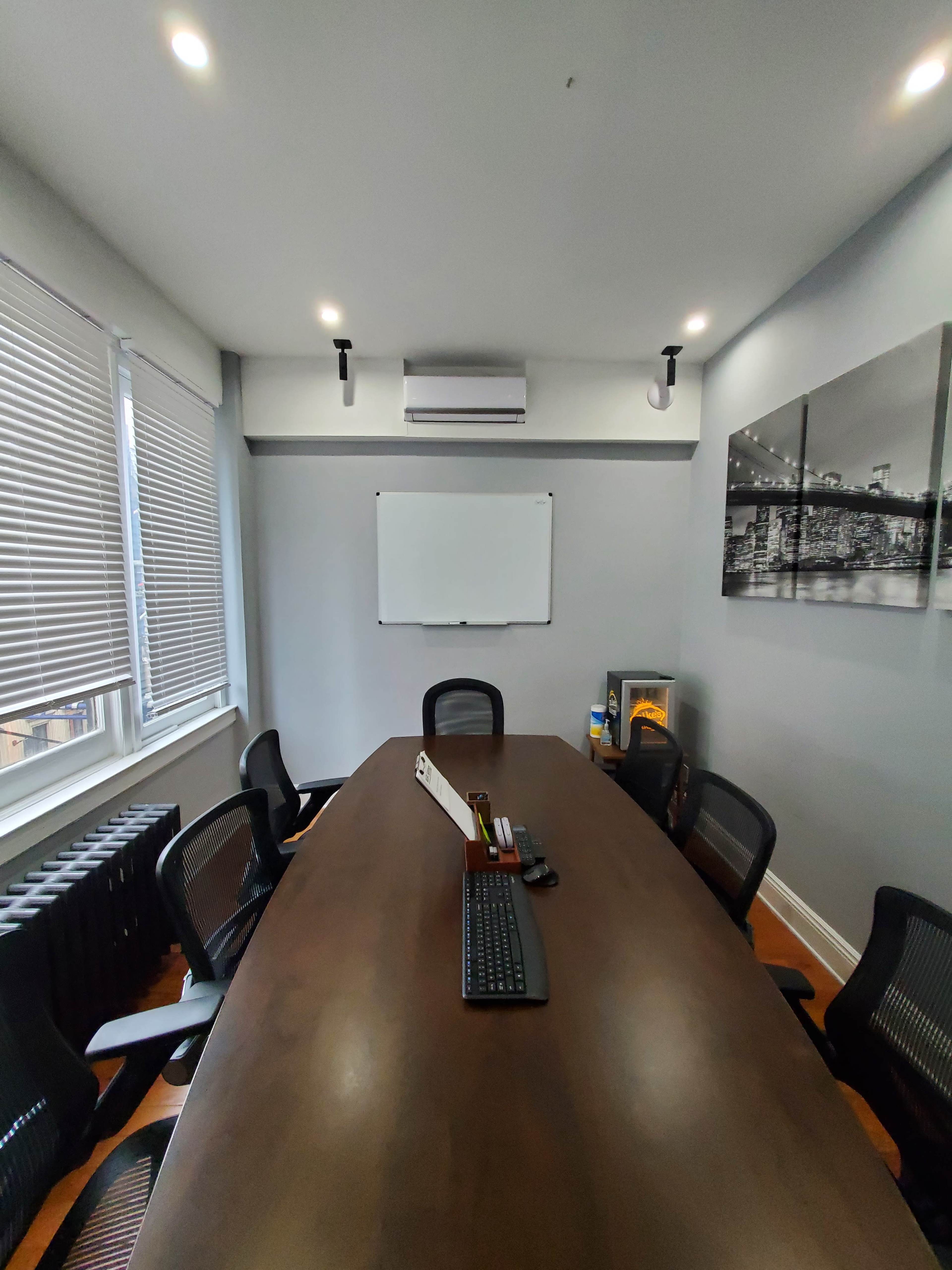 A conference room features a long wooden table surrounded by black mesh chairs, with a whiteboard and a wall-mounted air conditioner visible in the background.