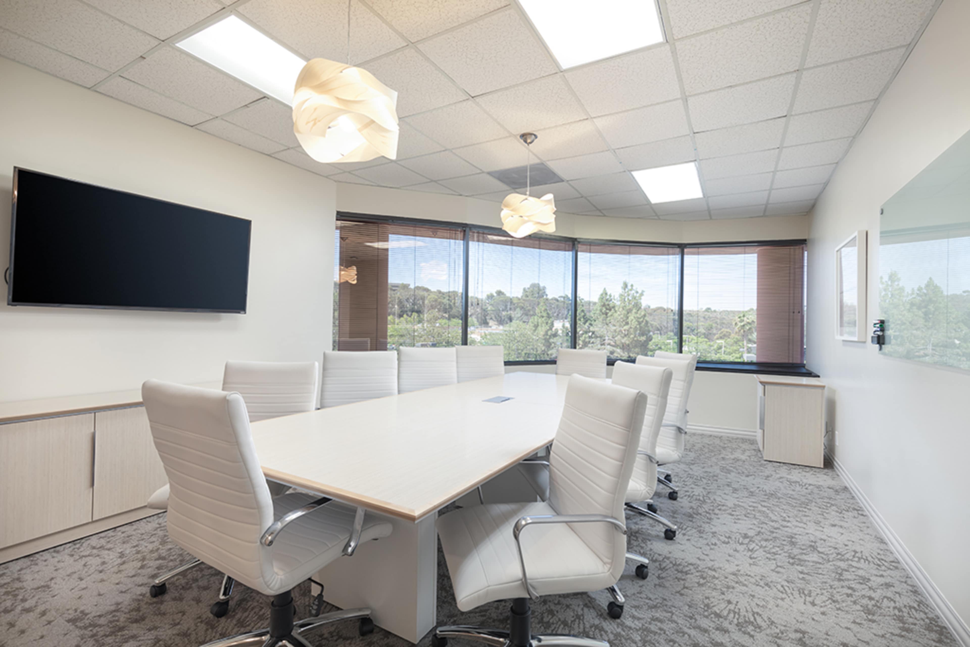 A modern conference room features a large table surrounded by white chairs, with a wall-mounted screen and windows overlooking greenery.