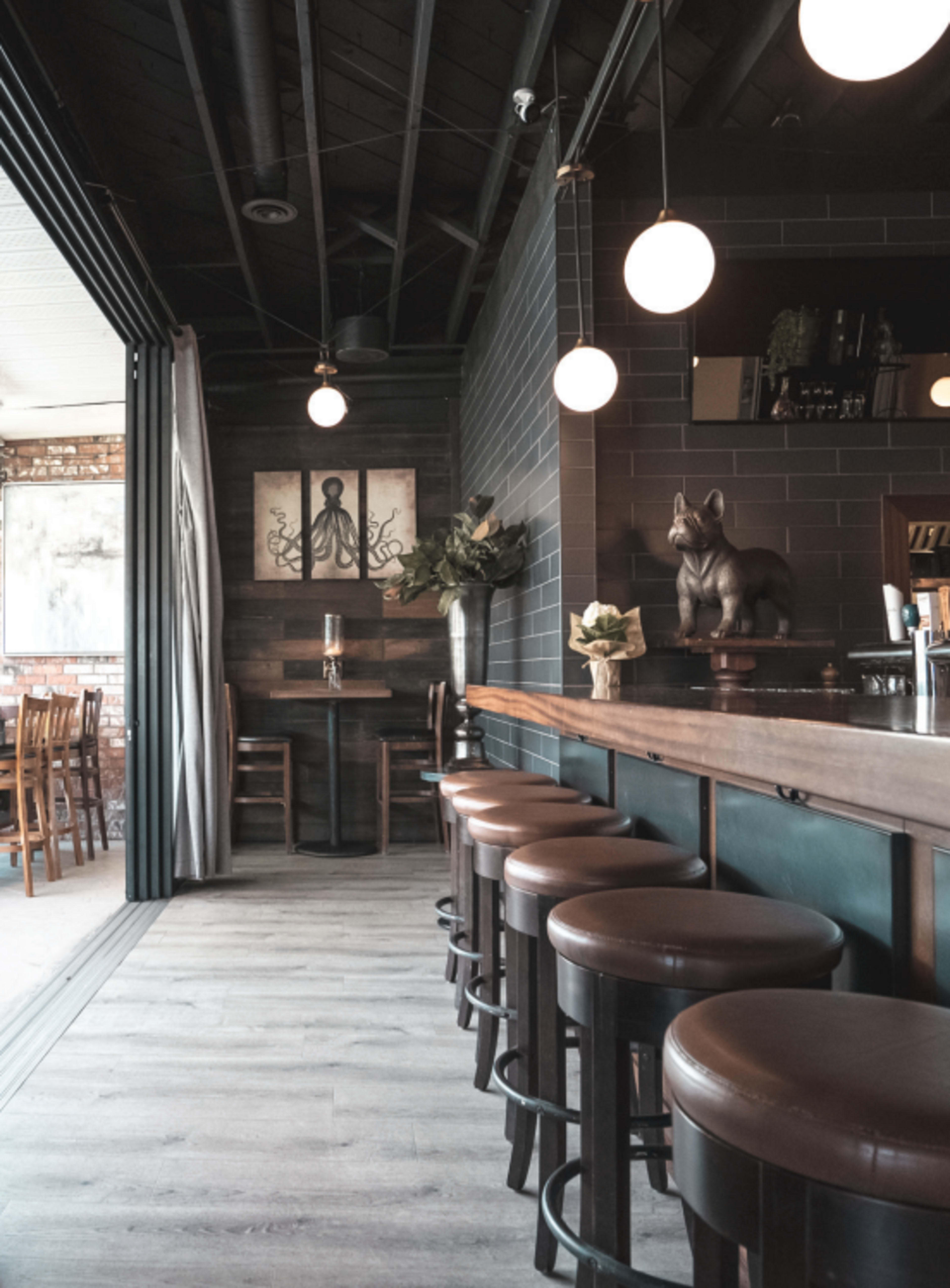 A modern bar area with wooden stools arranged along a long counter, featuring a dark color scheme and decorative elements like artwork and a statue.