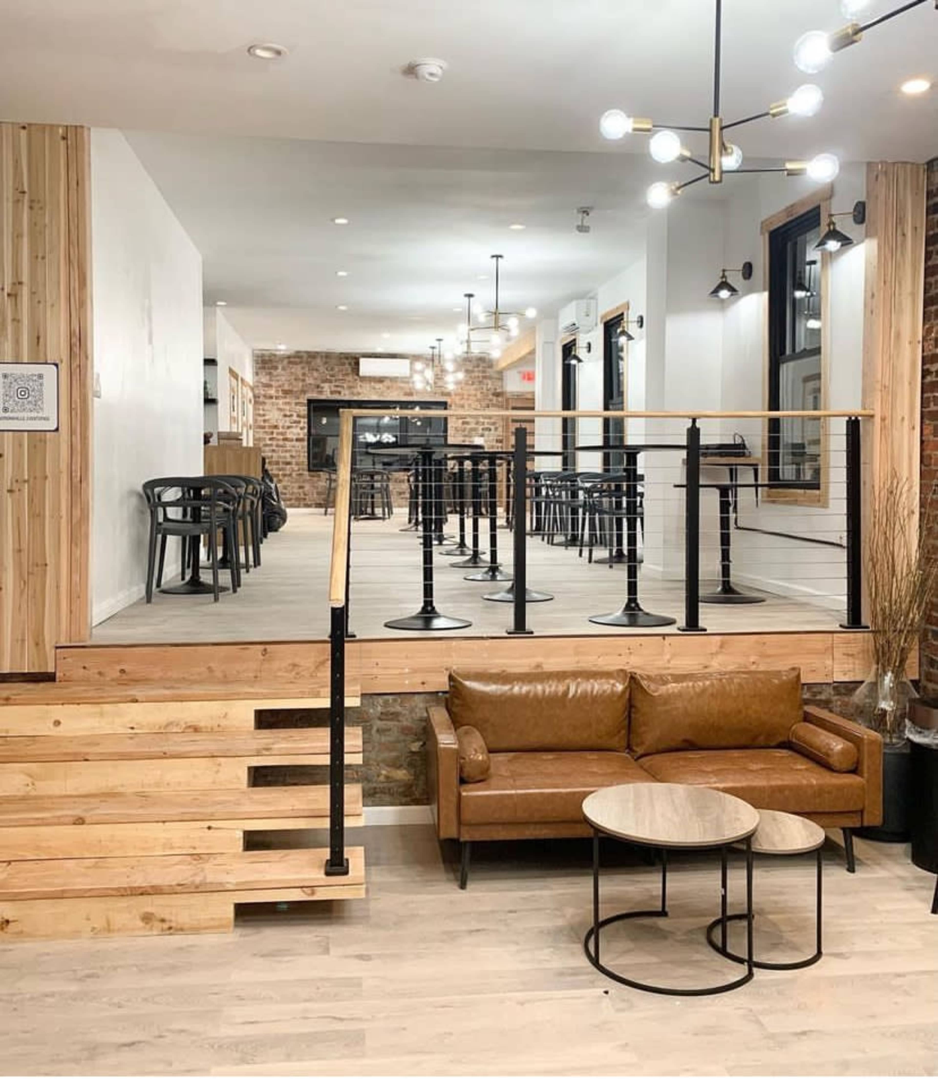 A modern café interior with wooden elements, a sunken seating area featuring a brown leather couch and coffee tables, and a bar-height seating area in the background.