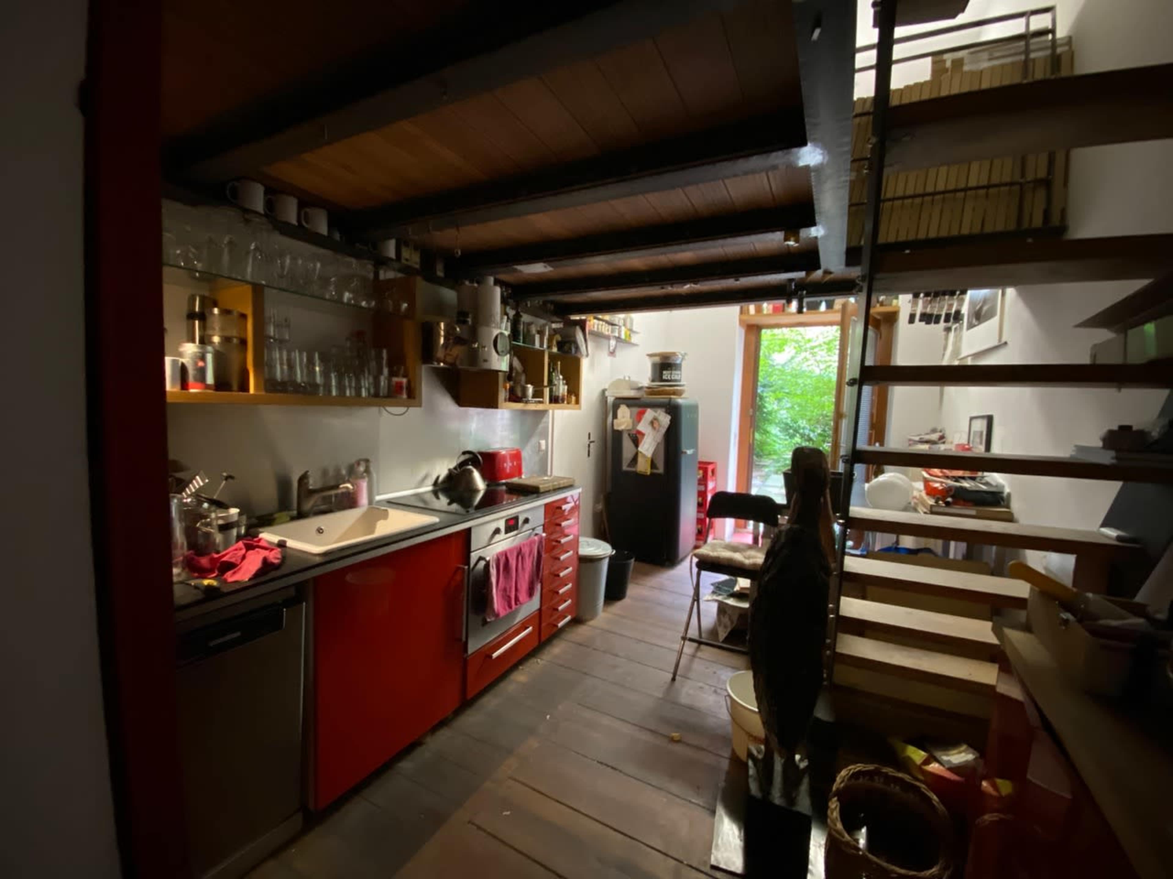 A compact kitchen with red cabinets, stainless steel appliances, and wooden beams, leading to a stairway and an open doorway filled with greenery.