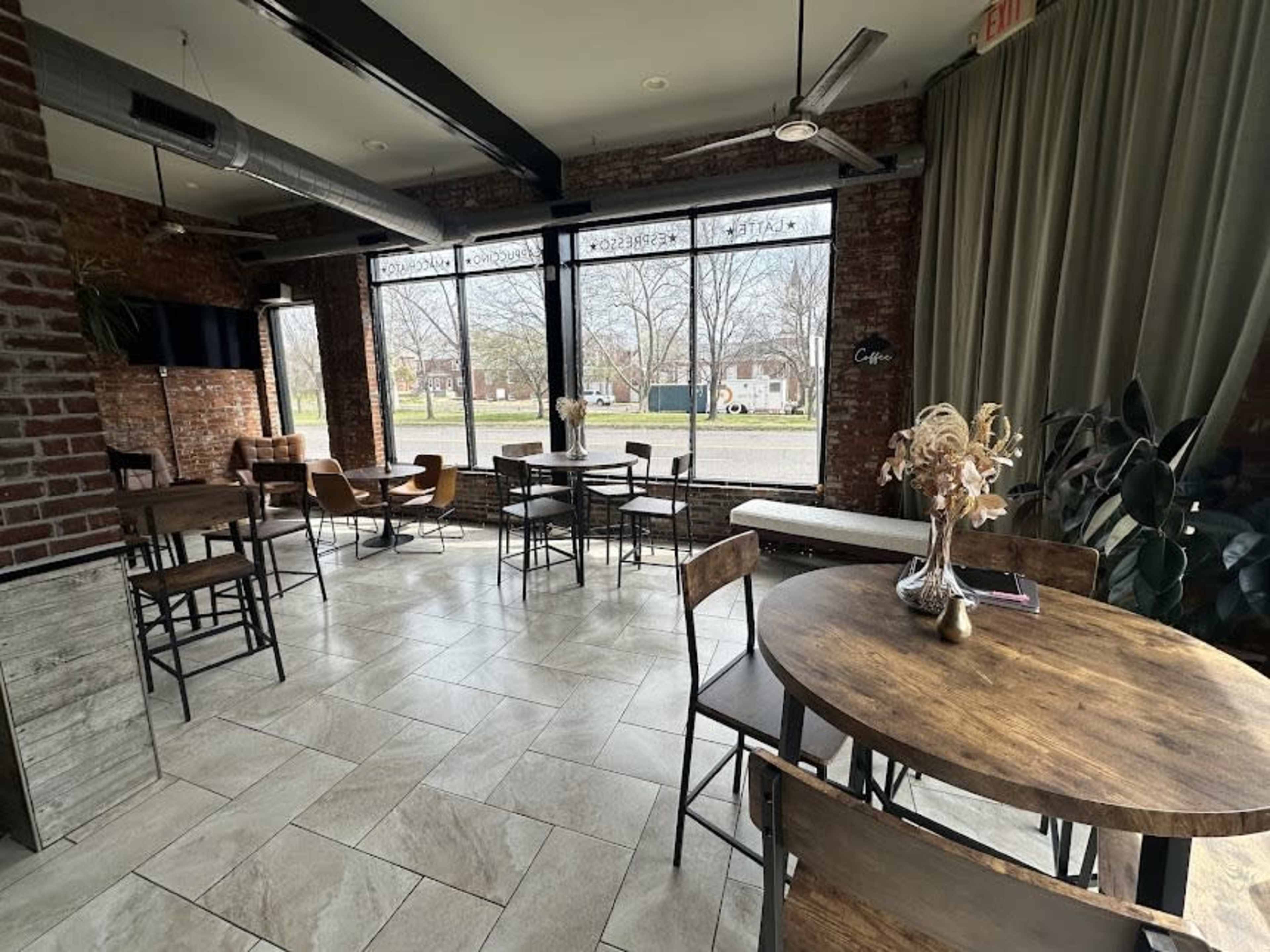 A cozy café interior features wooden tables and chairs, large windows with a view of the street, and exposed brick walls.
