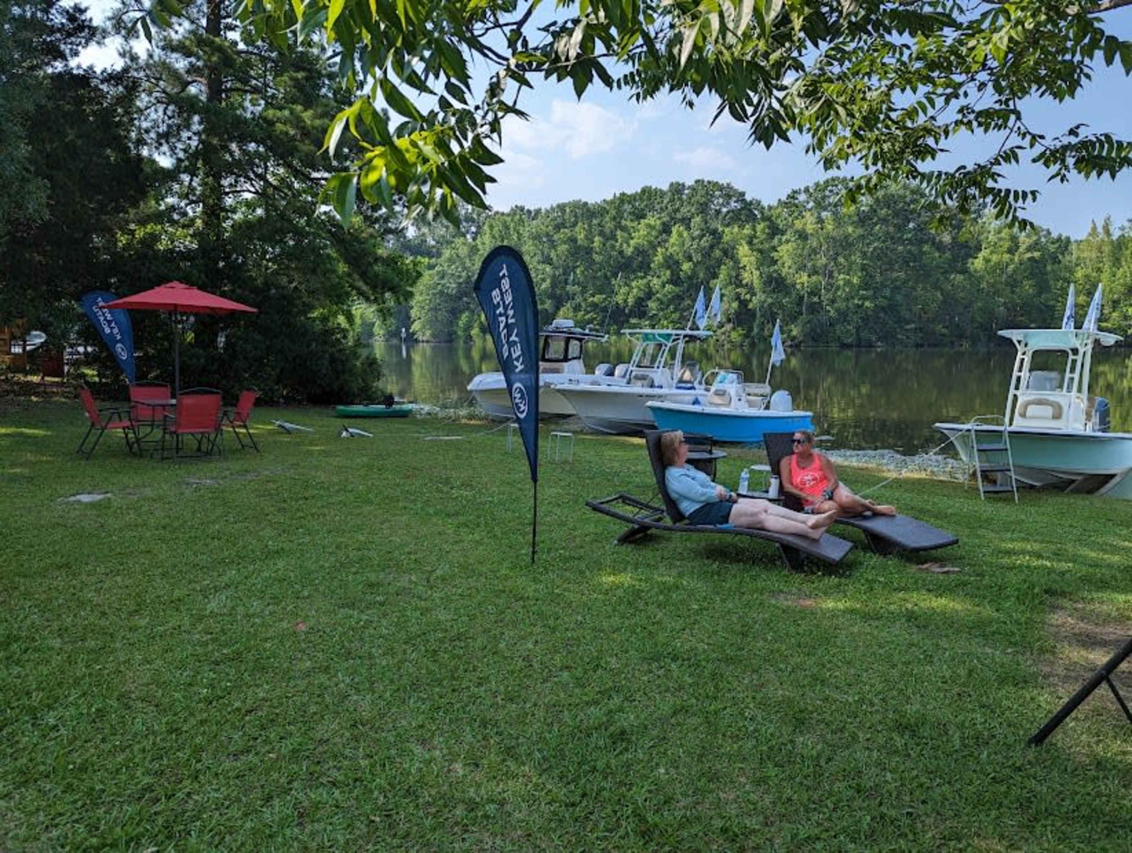 Two people relax on lounge chairs near a riverbank, with boats visible in the background and a shaded seating area set up nearby.