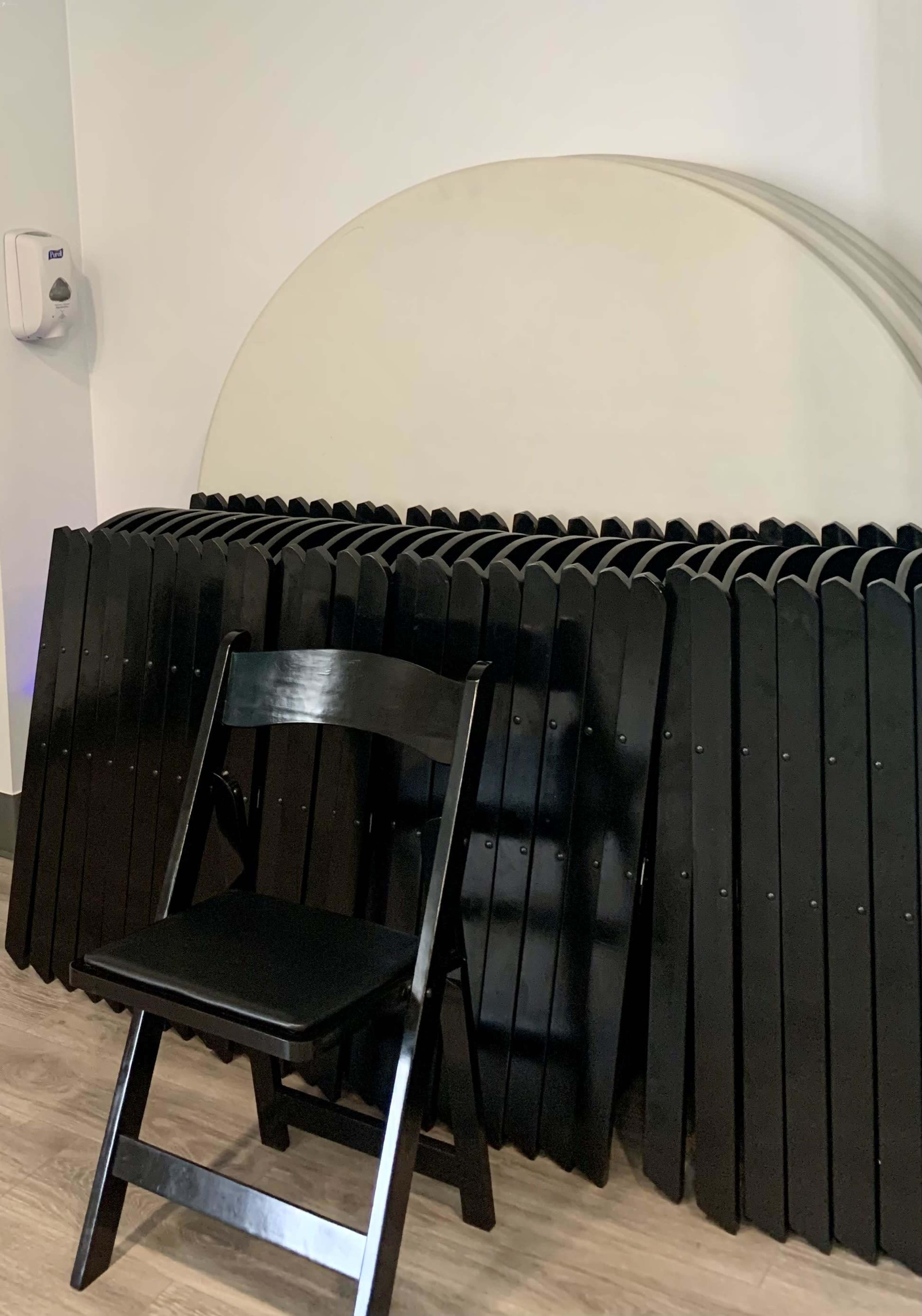A black folding chair sits in front of a neatly stacked row of black folding tables against a wall.